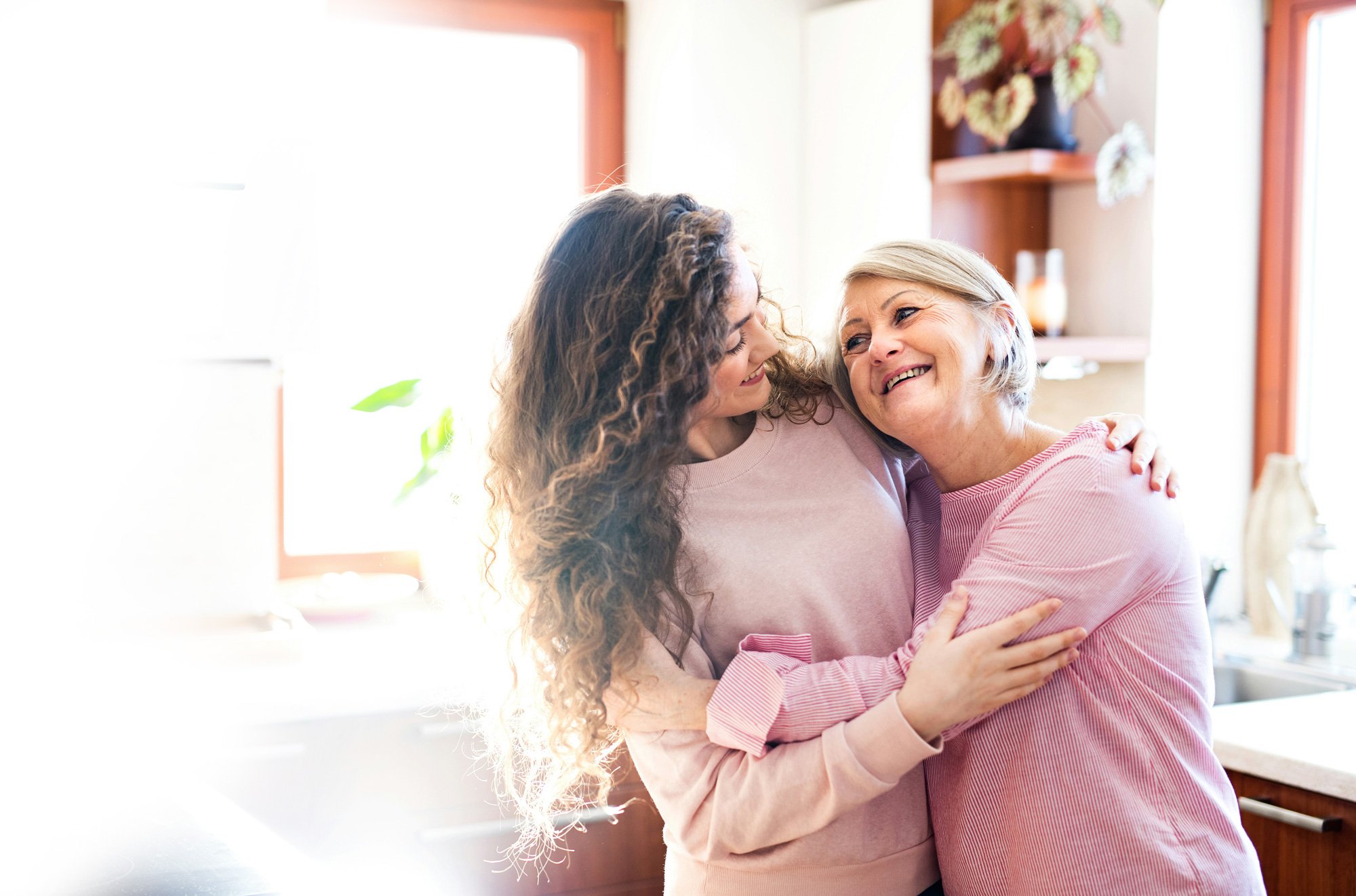 A young woman hugging an older woman in a bright kitchen, both smiling and looking happy.