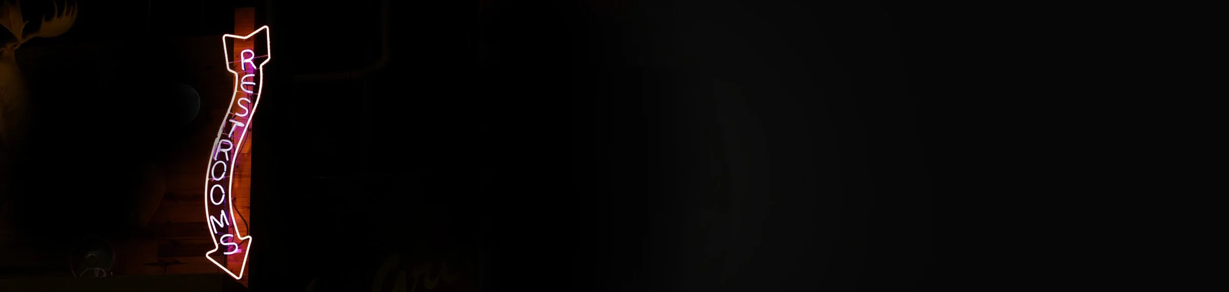 Vertical neon sign spelling 'ROOSTROOM'S' with an arrow pointing down, mounted on a wooden wall in a dimly lit environment.