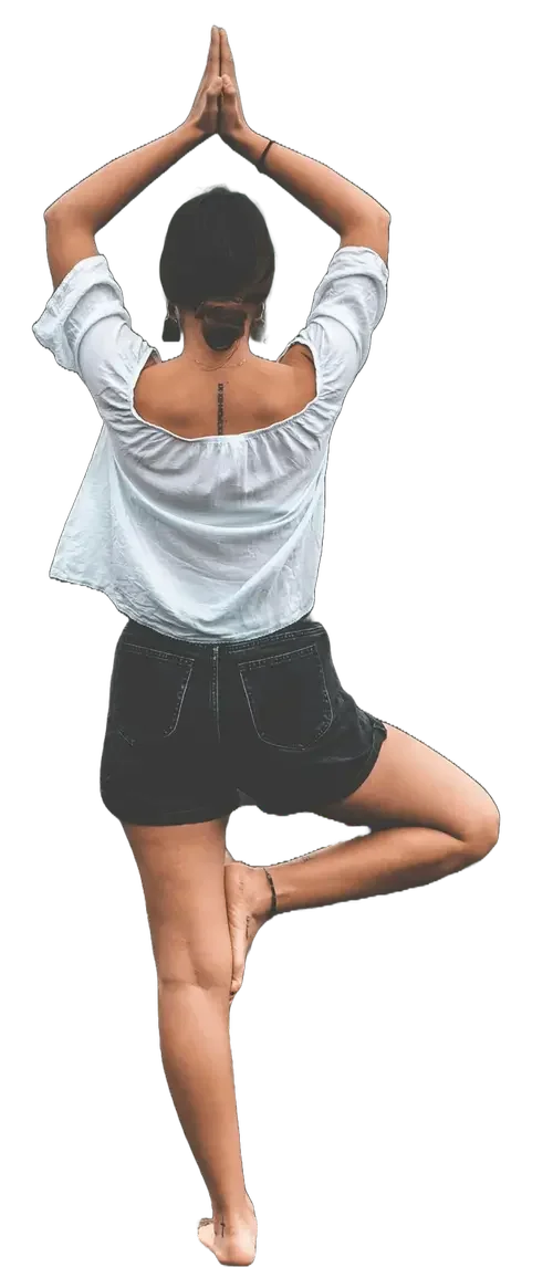 A woman practicing yoga or meditation, standing on one leg in a tree pose with hands in prayer position above her head, wearing a white blouse and black shorts, with her back to the camera