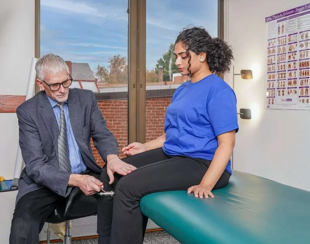 A healthcare professional, a woman in blue scrubs, examining a patient's knee in a medical office with an anatomy poster on the wall and a window showing rooftops outside.