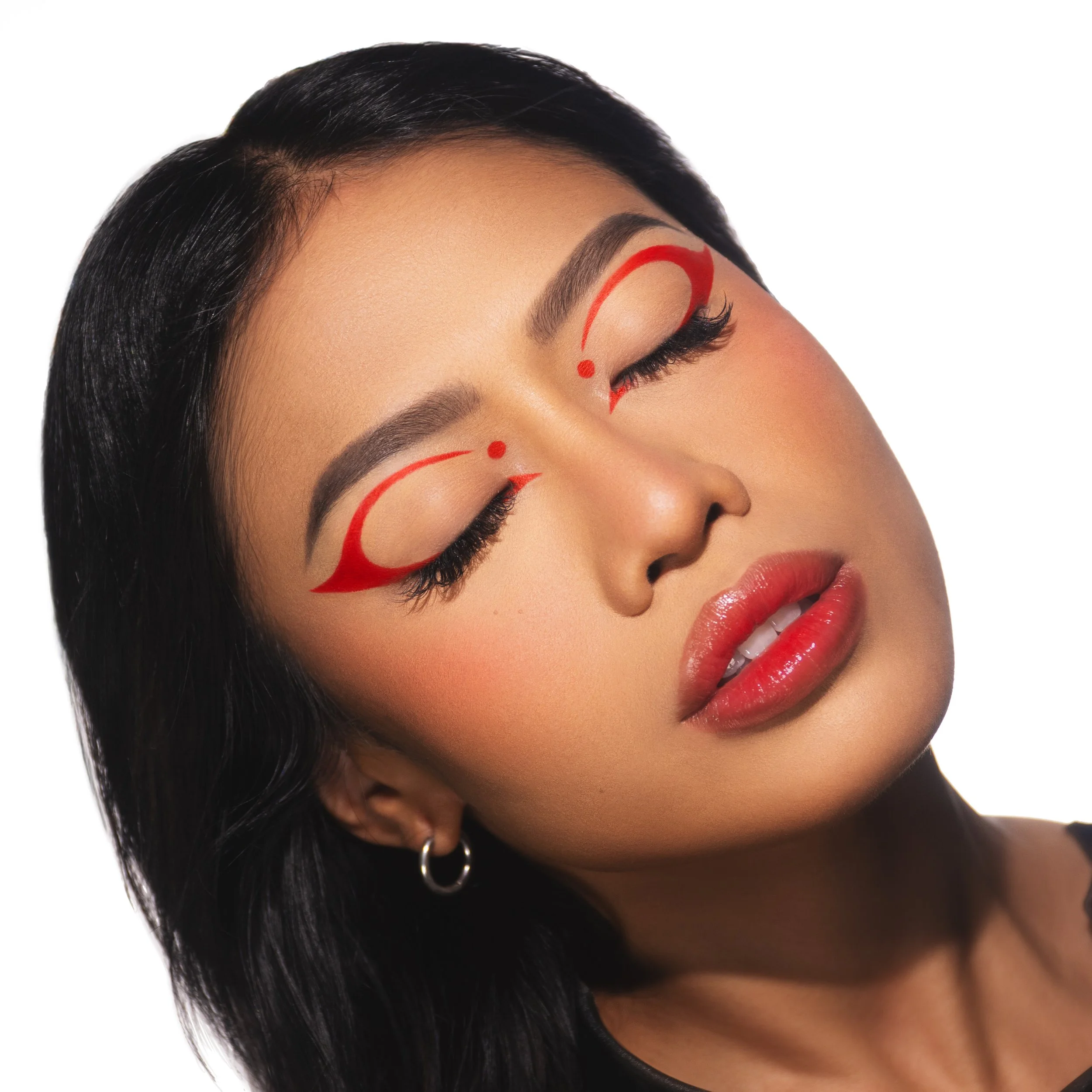 Close-up of a woman with dark hair and makeup, closed eyes, and red artistic face paint near her eyes.