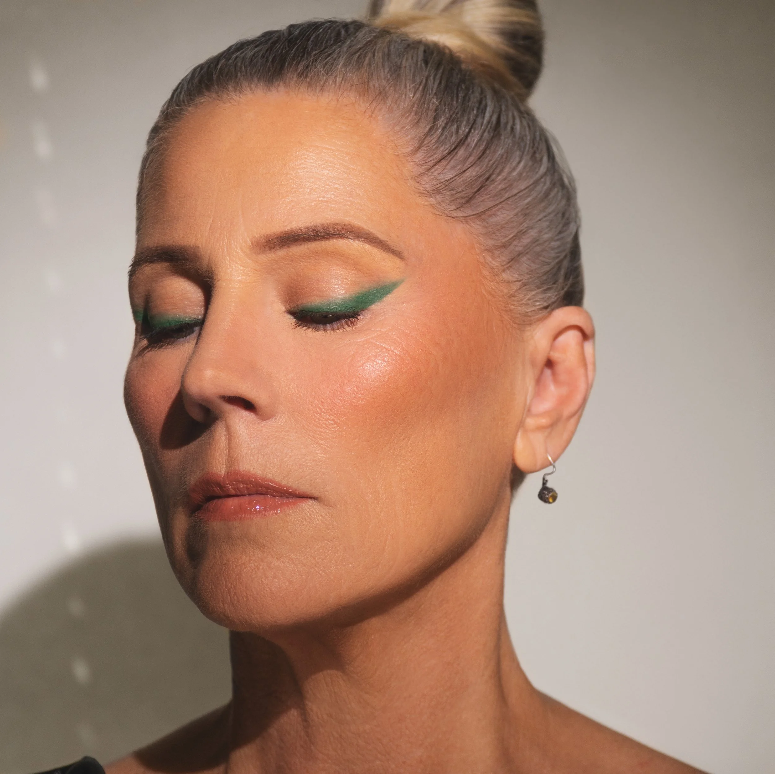 Close-up of a woman with her eyes closed, wearing green eyeliner, with her hair in a bun, and a single earring visible.