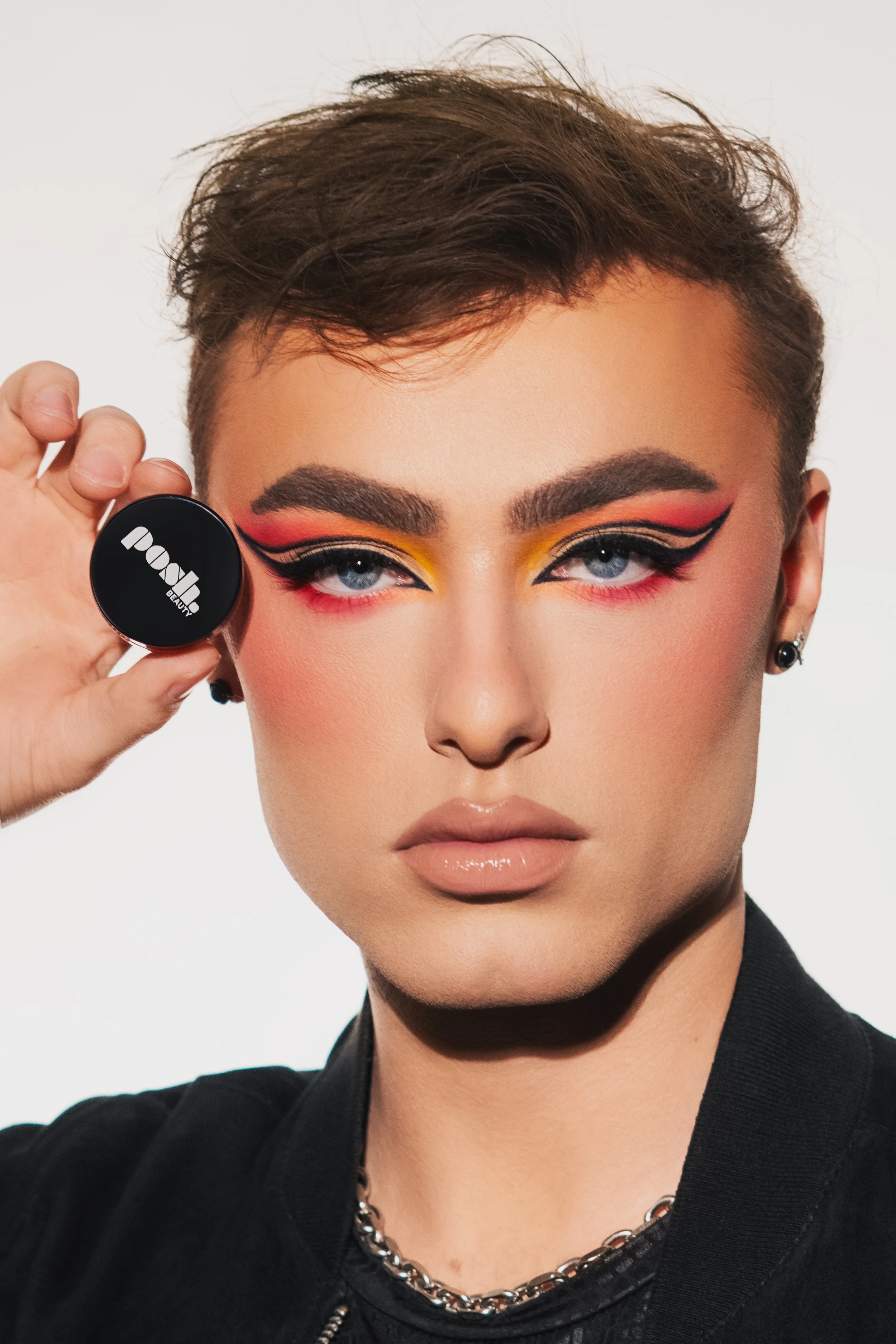 Person holding a black container of eyeshadow, with bold and colorful eye makeup, wearing a black top and a silver chain necklace.