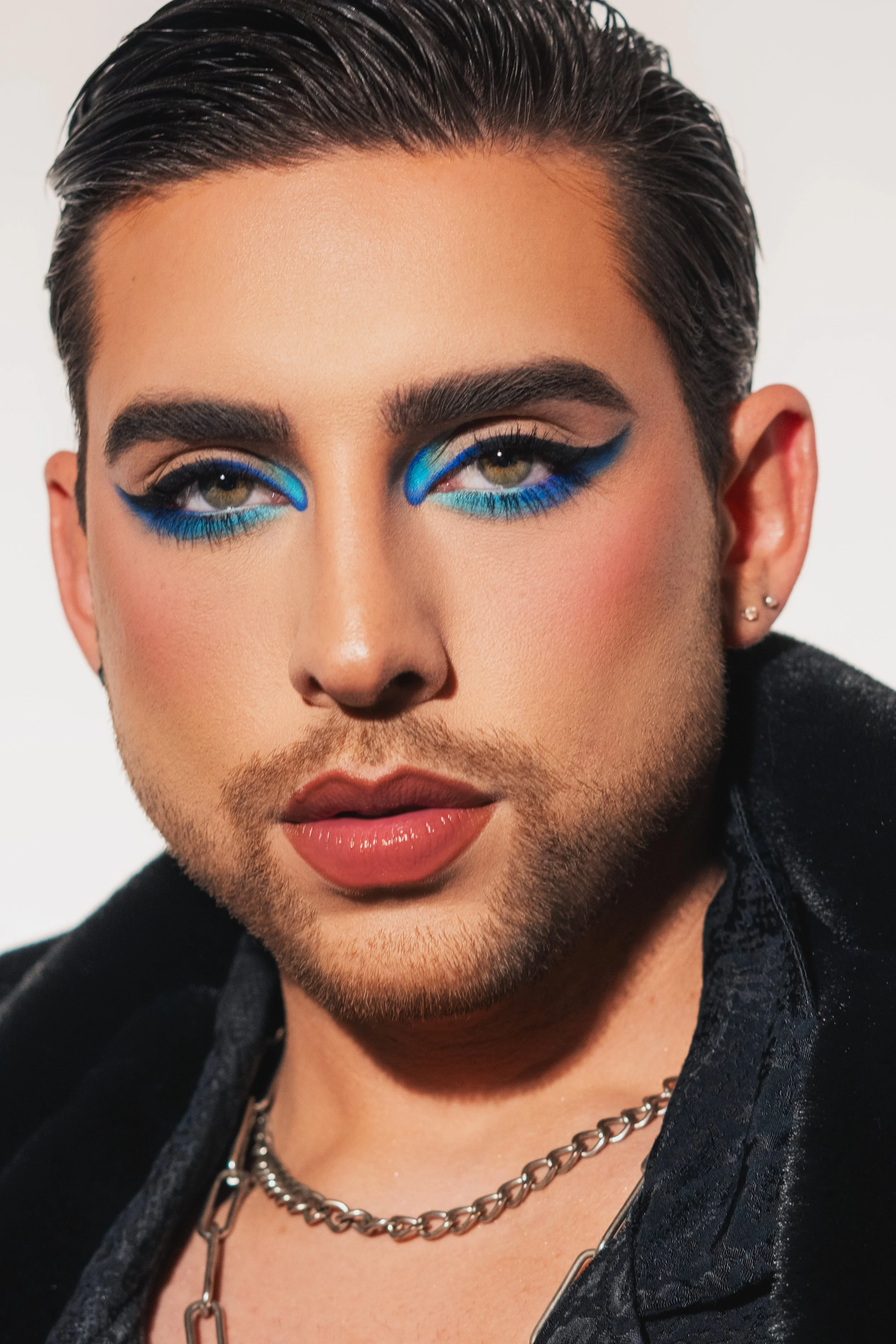 Close-up of a person with styled dark hair, bold makeup featuring bright blue eyeshadow, eyeliner, and glossy lips, wearing multiple earrings and silver chains, against a plain background.
