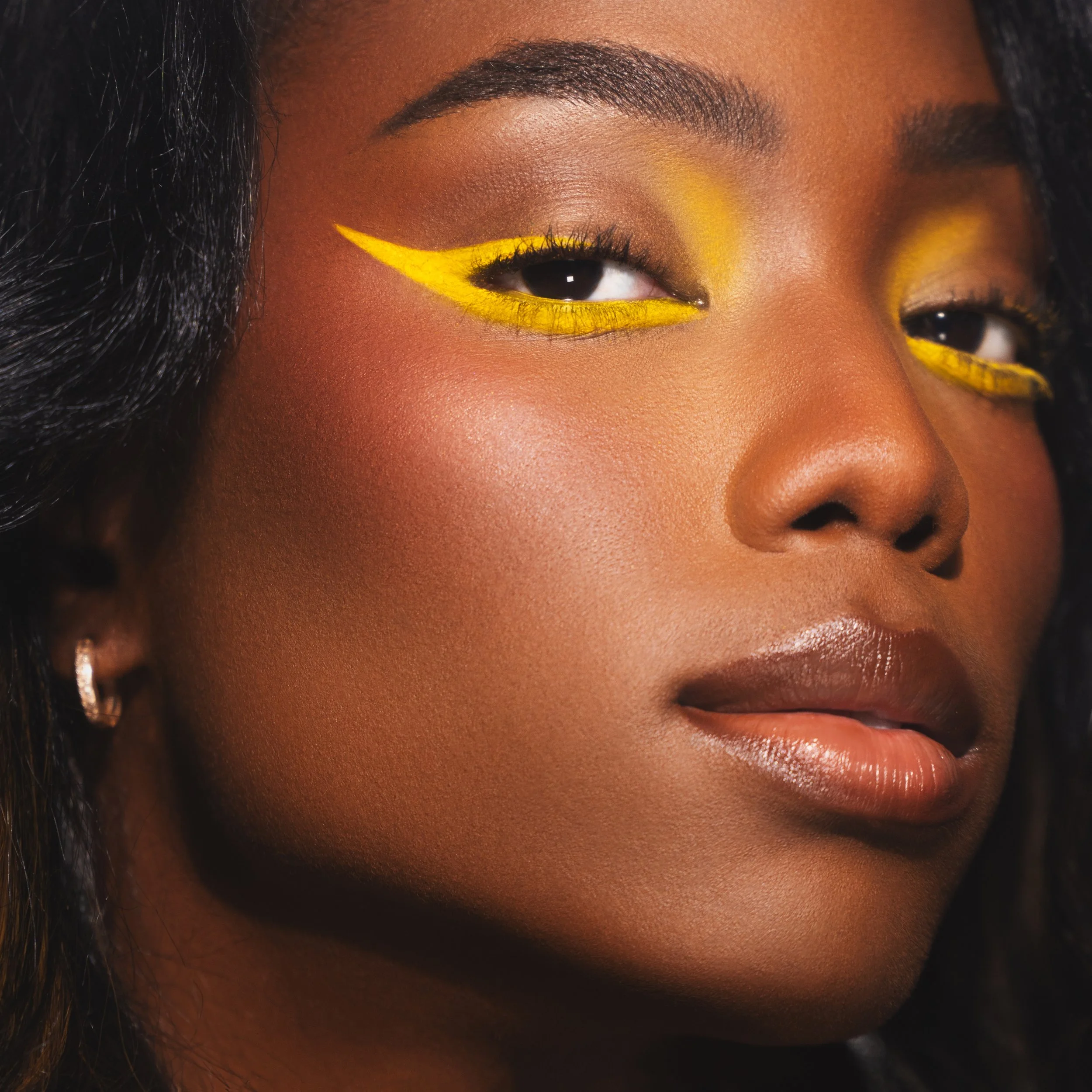 A close-up of a woman's face showing her eye with vibrant yellow eyeshadow with a winged eyeliner, full lips with glossy makeup, and smooth skin, with dark hair on the side.