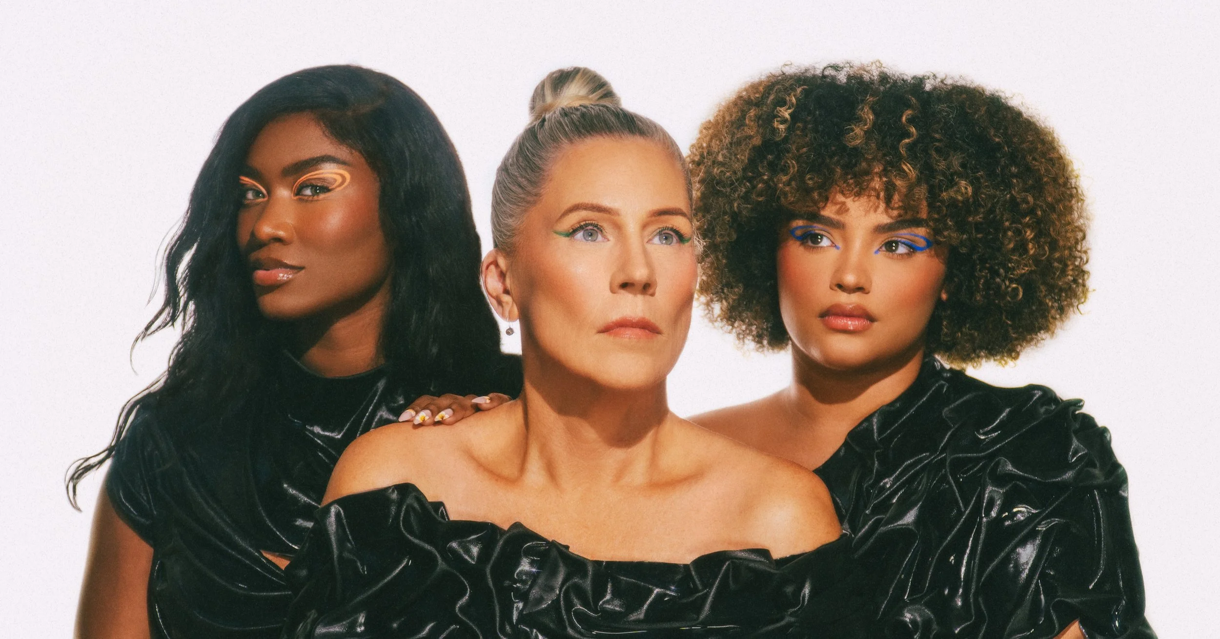 Three women with bold makeup posing together against a white background. The woman on the left has dark curly hair and gold eye makeup. The woman in the middle has blonde hair styled in a bun and green eyeliner. The woman on the right has voluminous