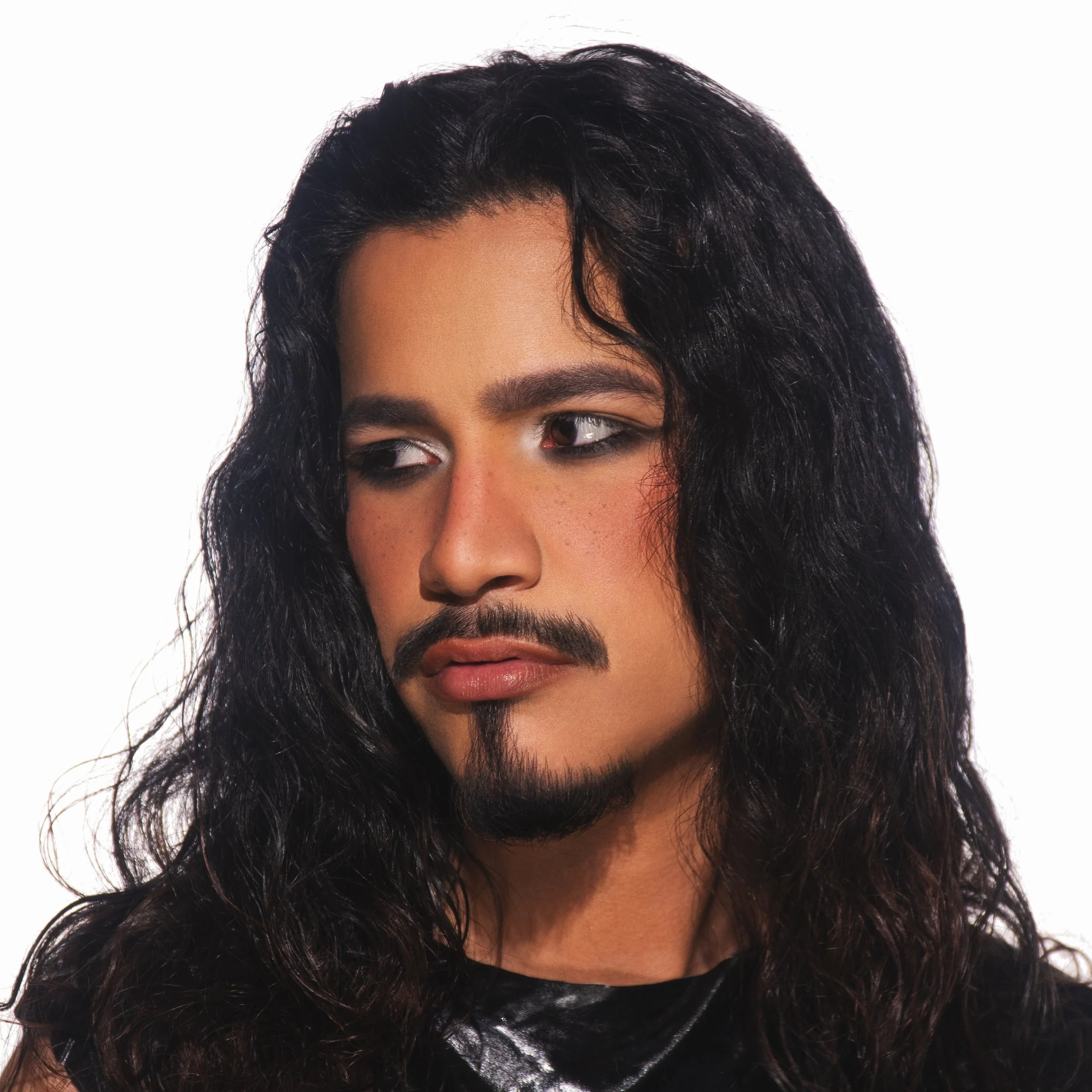 Close-up of a man with long, black, curly hair and a goatee, looking to the side, wearing a black shirt with a shiny fabric.