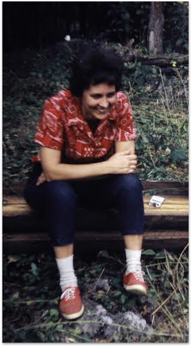 A person with short dark hair smiling, dressed in a red patterned shirt, black pants, white socks, and red sneakers, sitting on a wooden log in a forested area with greenery around.