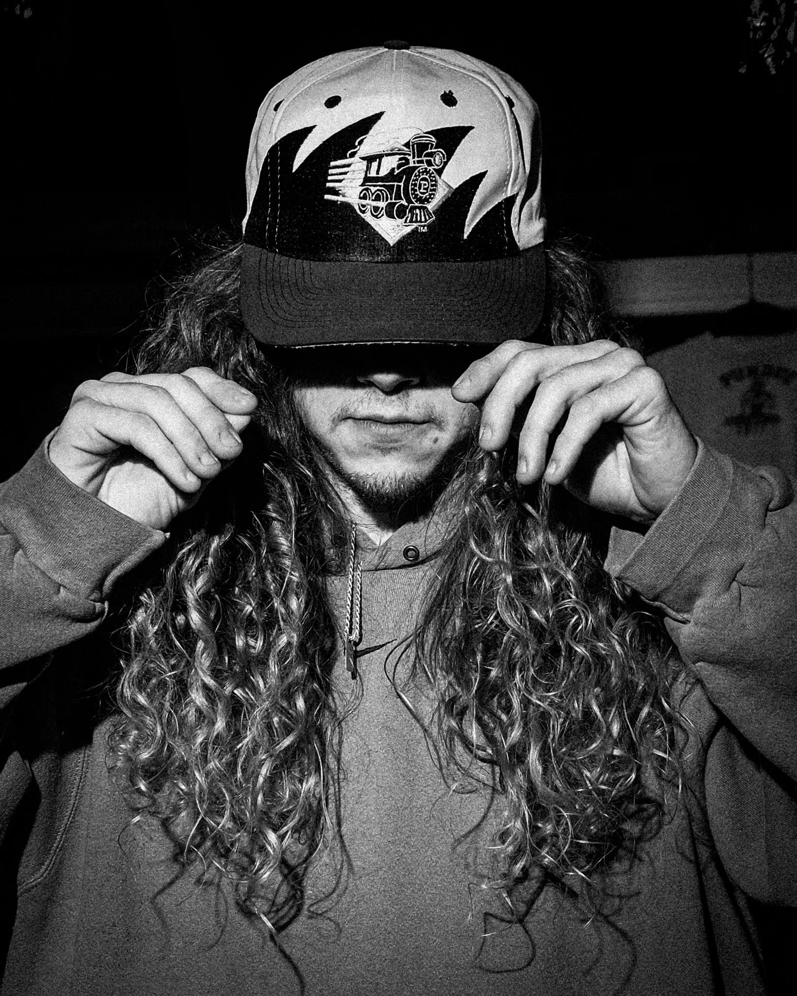 A person with long, curly hair is wearing a baseball cap featuring a train illustration. The individual is holding the brim of the cap with both hands, partially covering their face, which has a light beard and mustache. The person is dressed in a ho