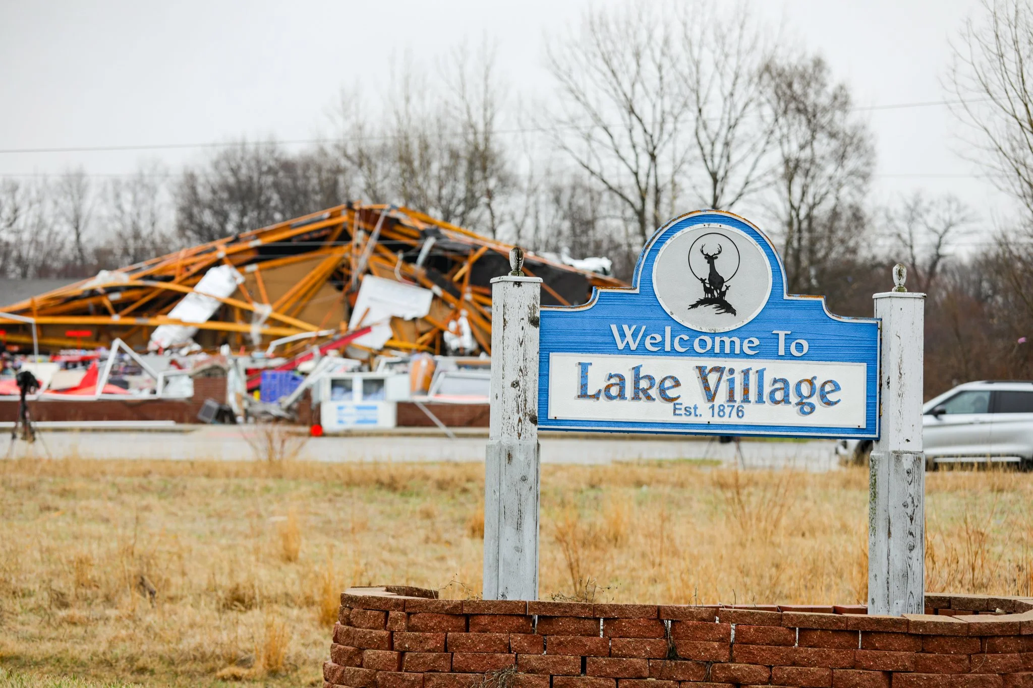 Overturned boat and debris behind a blue welcome sign for Lake Village, established in 1876, with a cluster of leafless trees in the background.