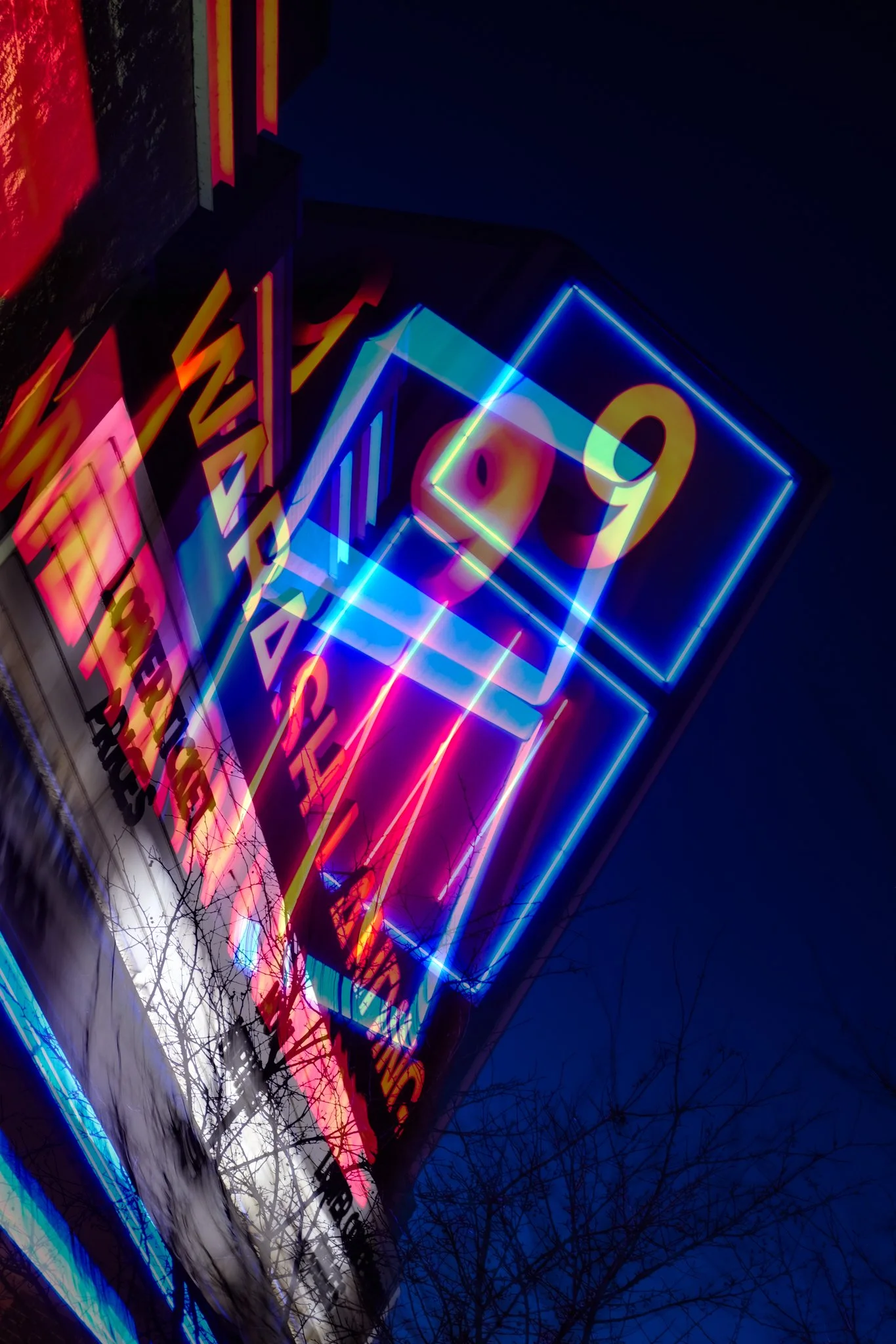 Colorful neon signs with various shapes and words, displayed on the side of a building at night, with bare tree branches in the foreground.