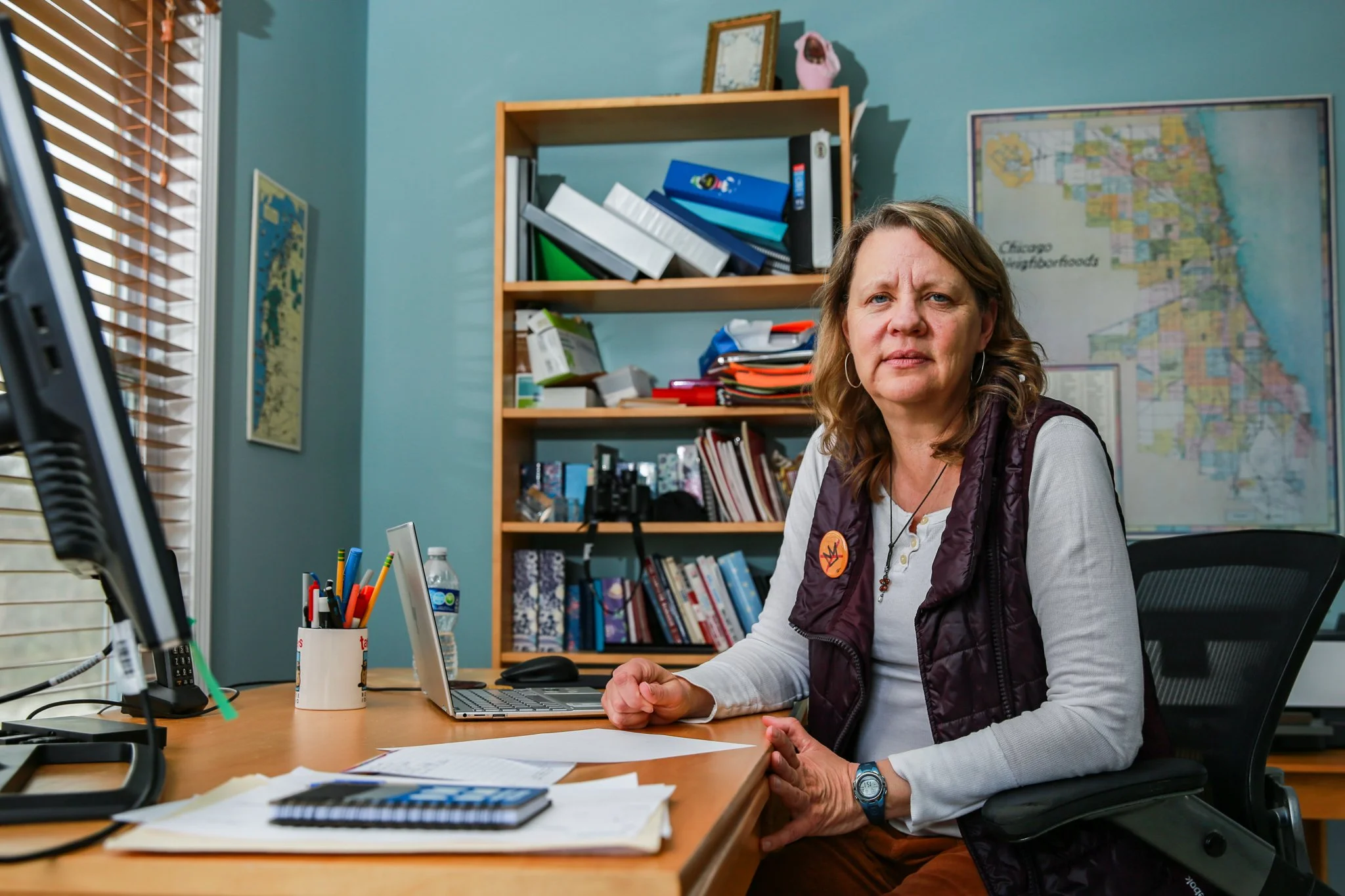 A woman with shoulder-length hair sitting at an office desk, wearing a white shirt and a purple vest, with papers, a laptop, and office supplies on the desk, in a room with a bookshelf, a map on the wall, and a large window with blinds.