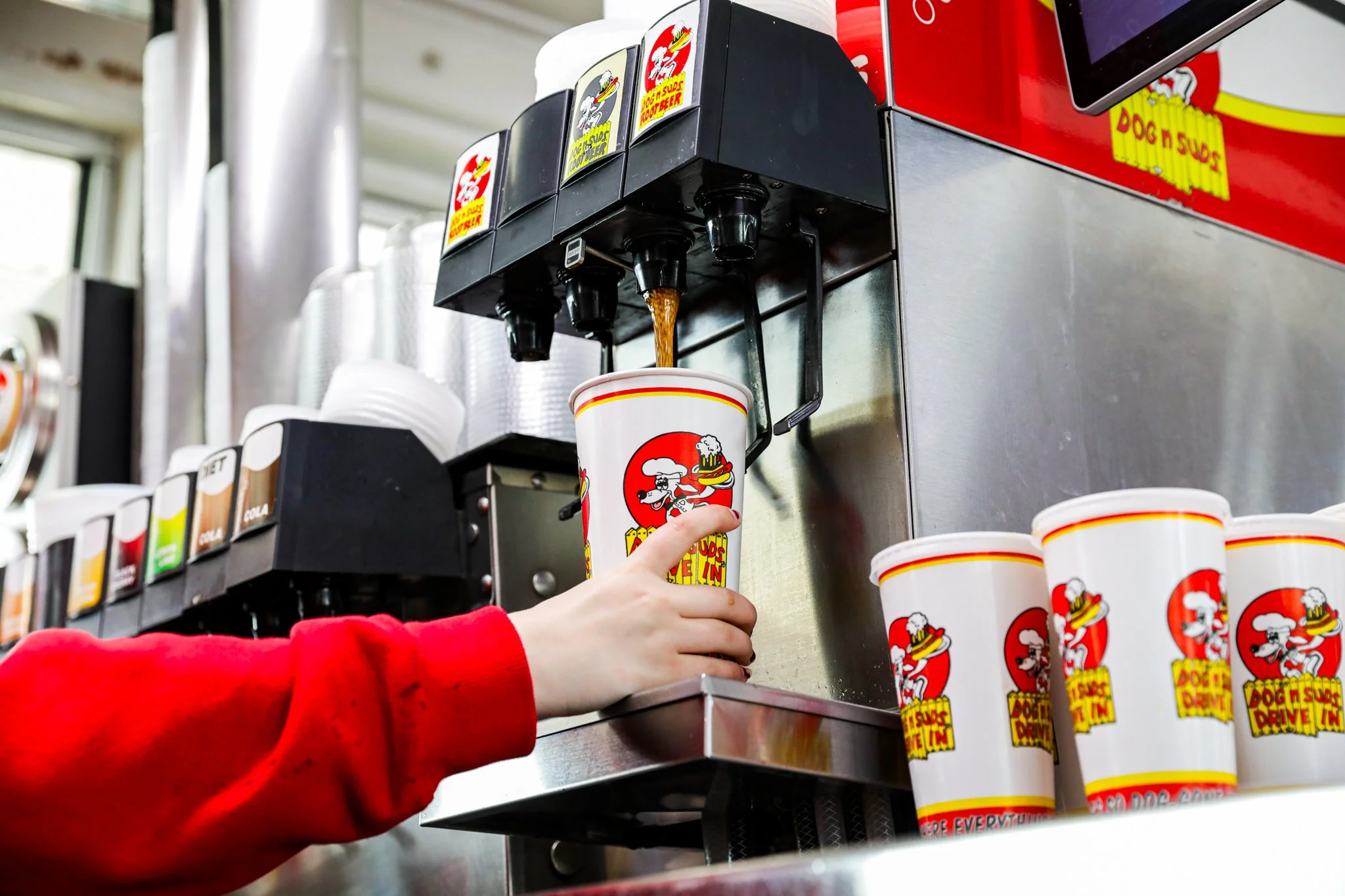 A person in a red long sleeve shirt is pressing an automatic soda dispenser to pour a soft drink into a cup with a dog and pizza logo. There are multiple cups with the logo stacked in front of the dispenser, and a row of soda flavor buttons.