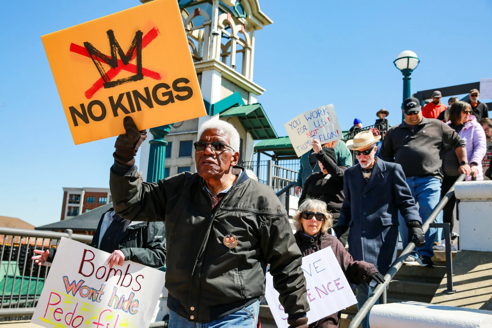 A group of protesters holding signs at an outdoor rally. One prominent sign reads 'NO KINGS' with a red strike through the letter 'M'. Other signs have various messages, and the crowd is diverse, walking down stairs with a blue sky in the background.