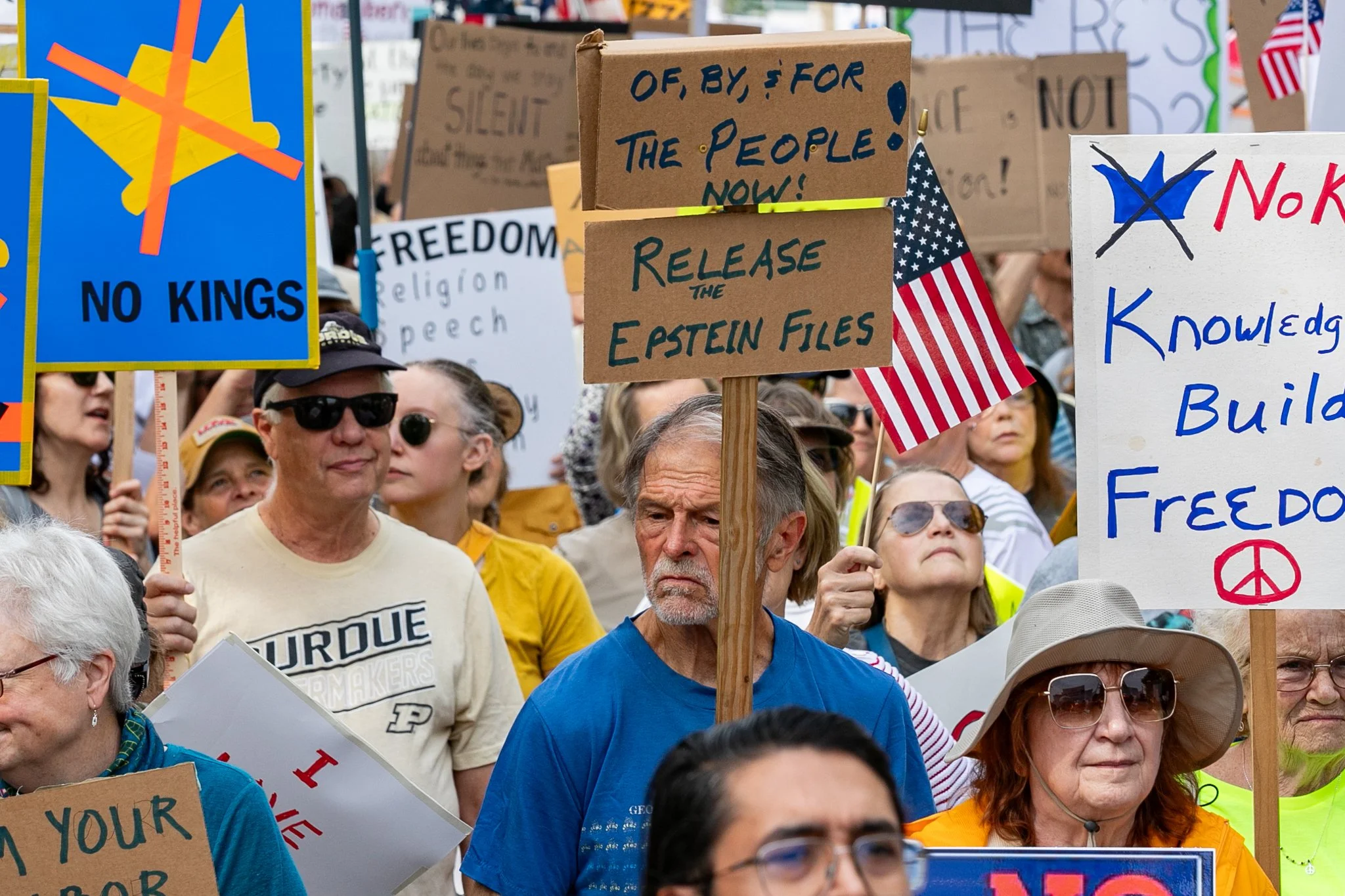 Crowd of people at a protest or demonstration holding various signs, including one that says 'NO KINGS', and others with messages about freedom, the Epstein files, and government accountability. Some people are wearing sunglasses and hats, and there 