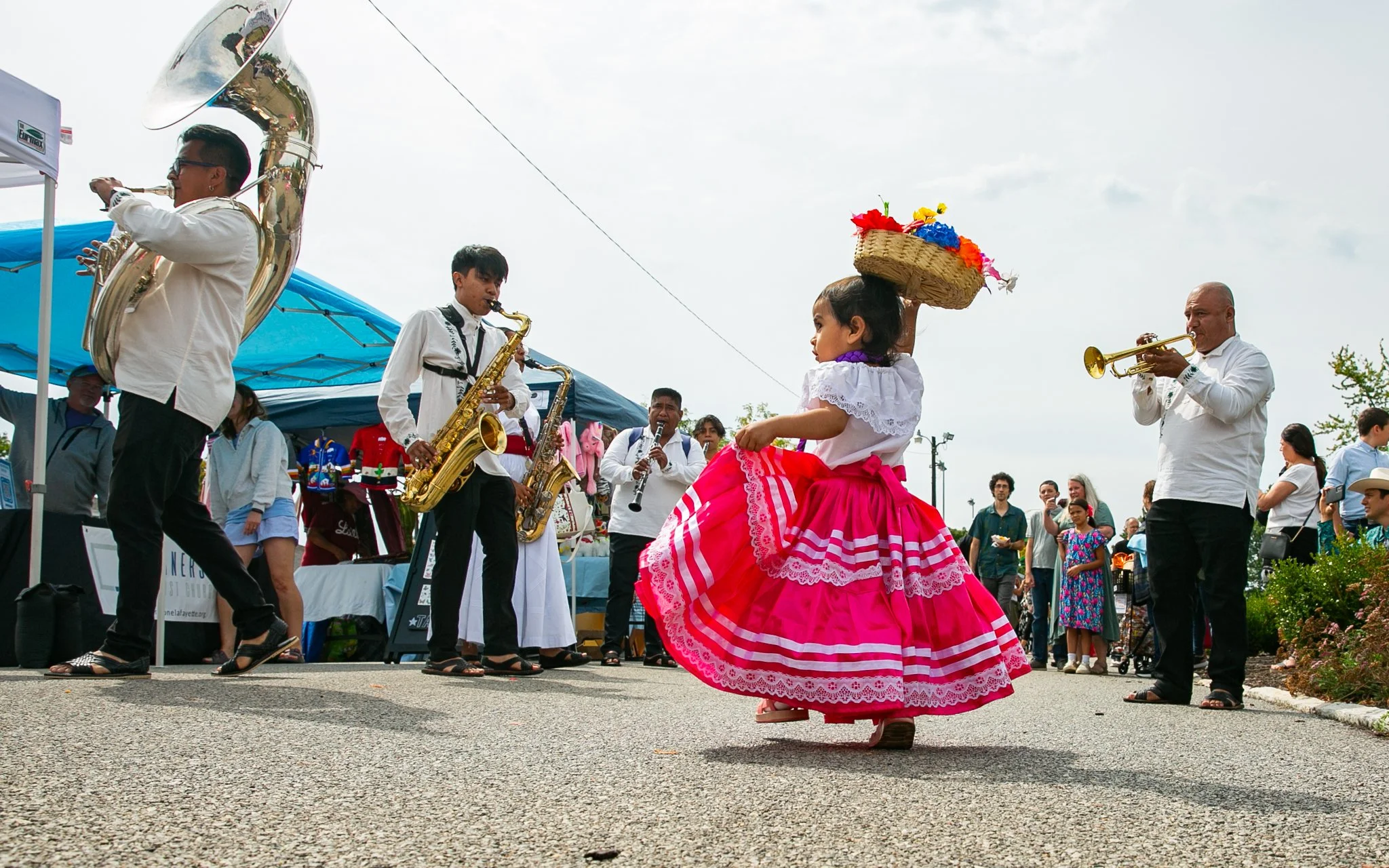 A young girl in a traditional dress with a pink skirt and lace details dances in front of a group playing musical instruments at an outdoor festival. The musicians are playing saxophones, trumpet, and clarinet, while a woman in the background wears a