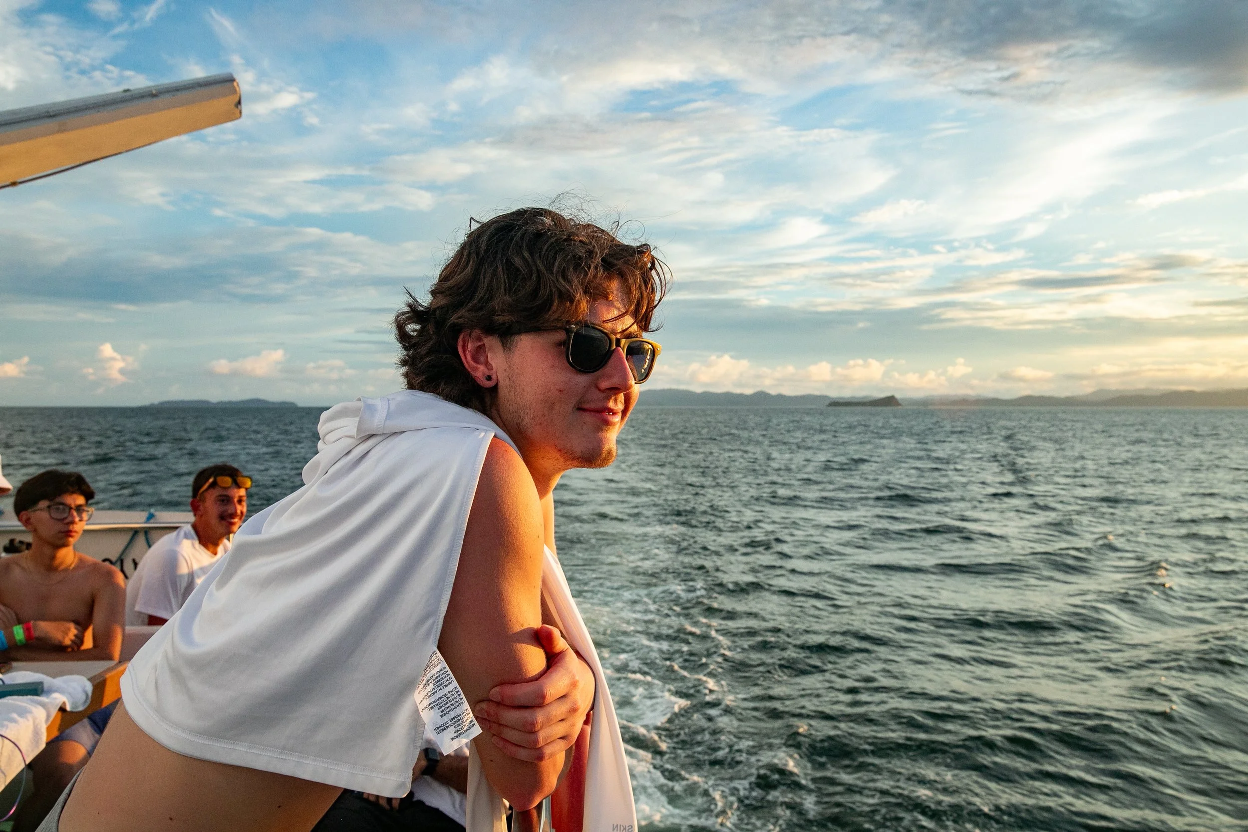 Young man with curly hair and sunglasses relaxing on a boat with friends, looking out over the ocean during sunset.