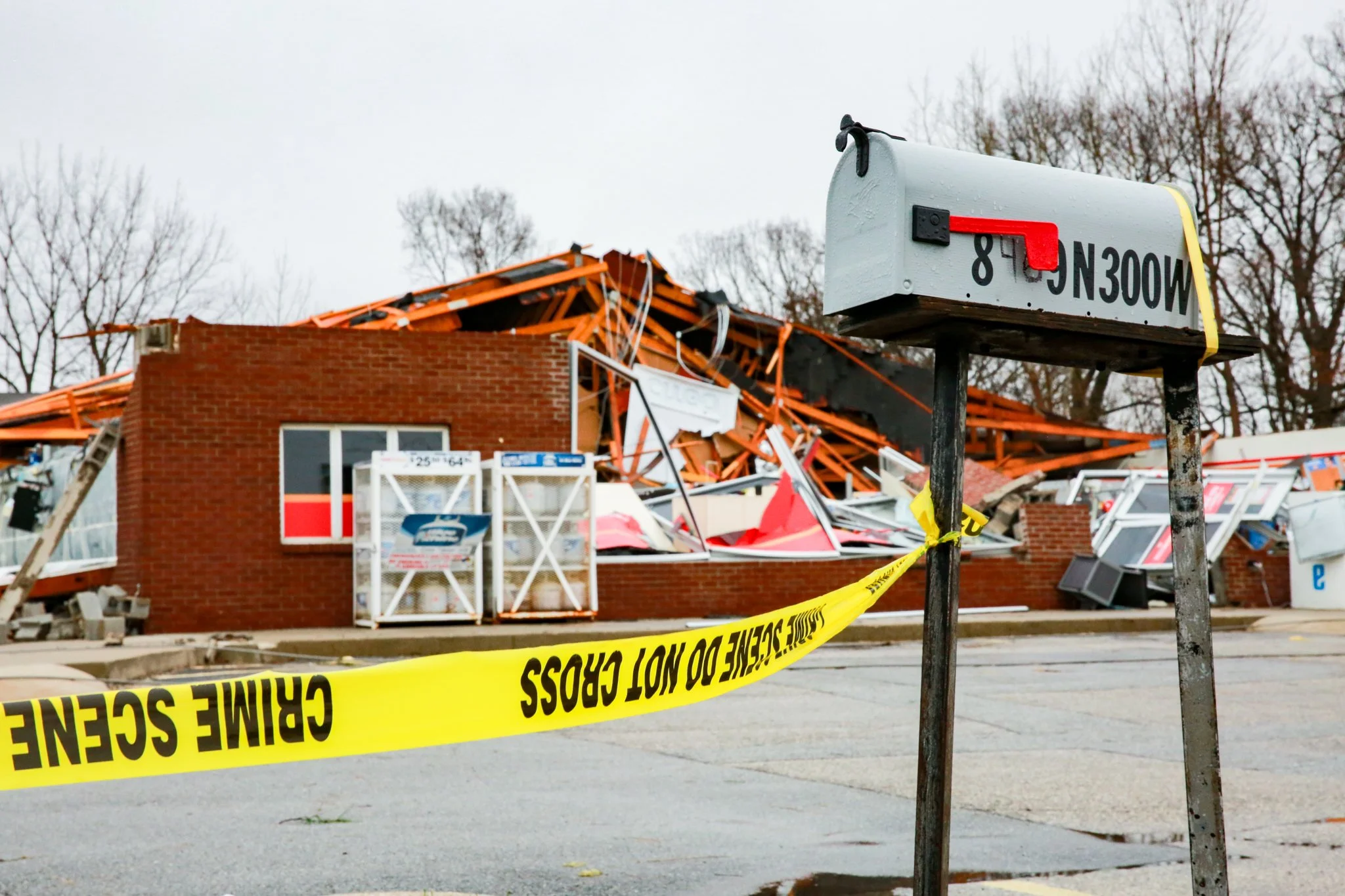 Collapsed building with orange framing and debris, surrounded by yellow caution tape that reads 'CRIME SCENE DO NOT CROSS', and a white signpost with partially visible black text.