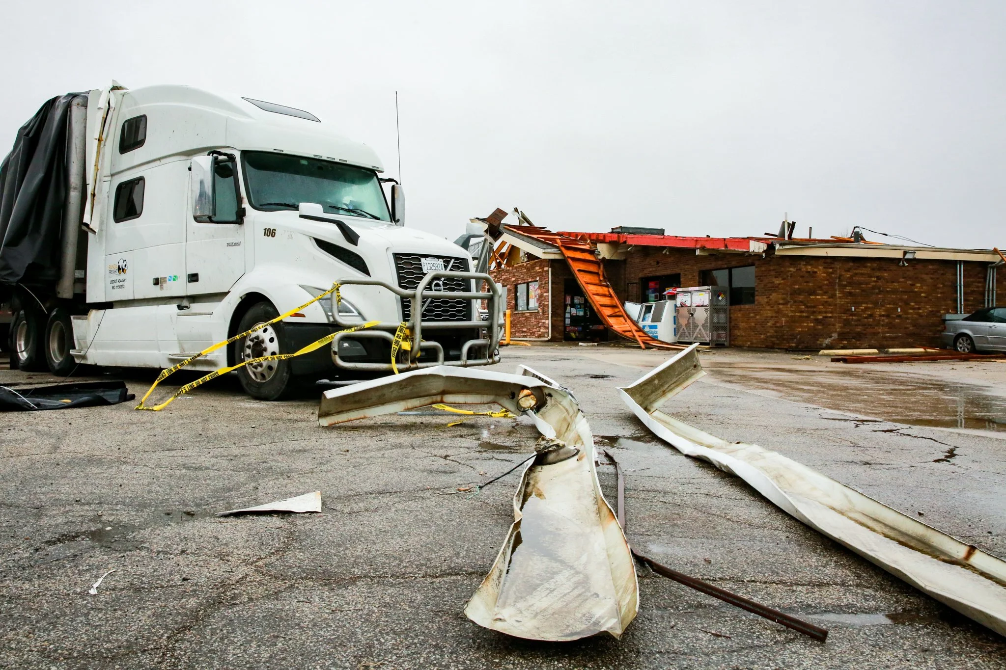 Debris and damage from a storm or accident at a building with a damaged roof, a large truck with crash barriers in front, and caution tape around the area.