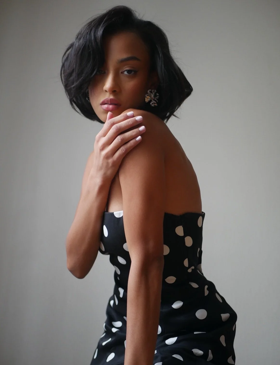 A woman with dark, shoulder-length hair, wearing a black dress with white polka dots, posing with her hand on her shoulder and looking at the camera.