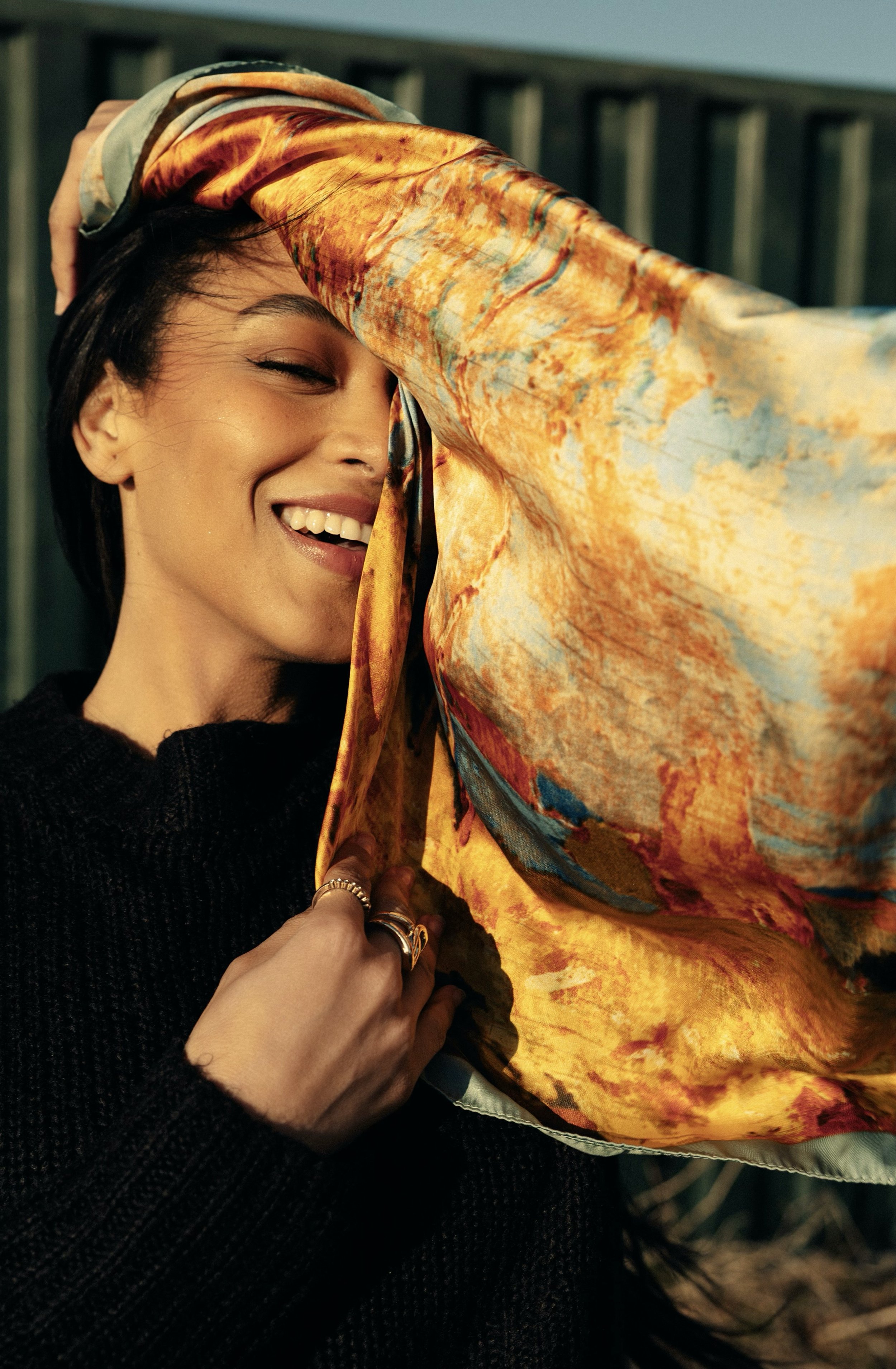 A woman smiling with her eyes closed, wearing a black sweater, holding a colorful scarf over her head.