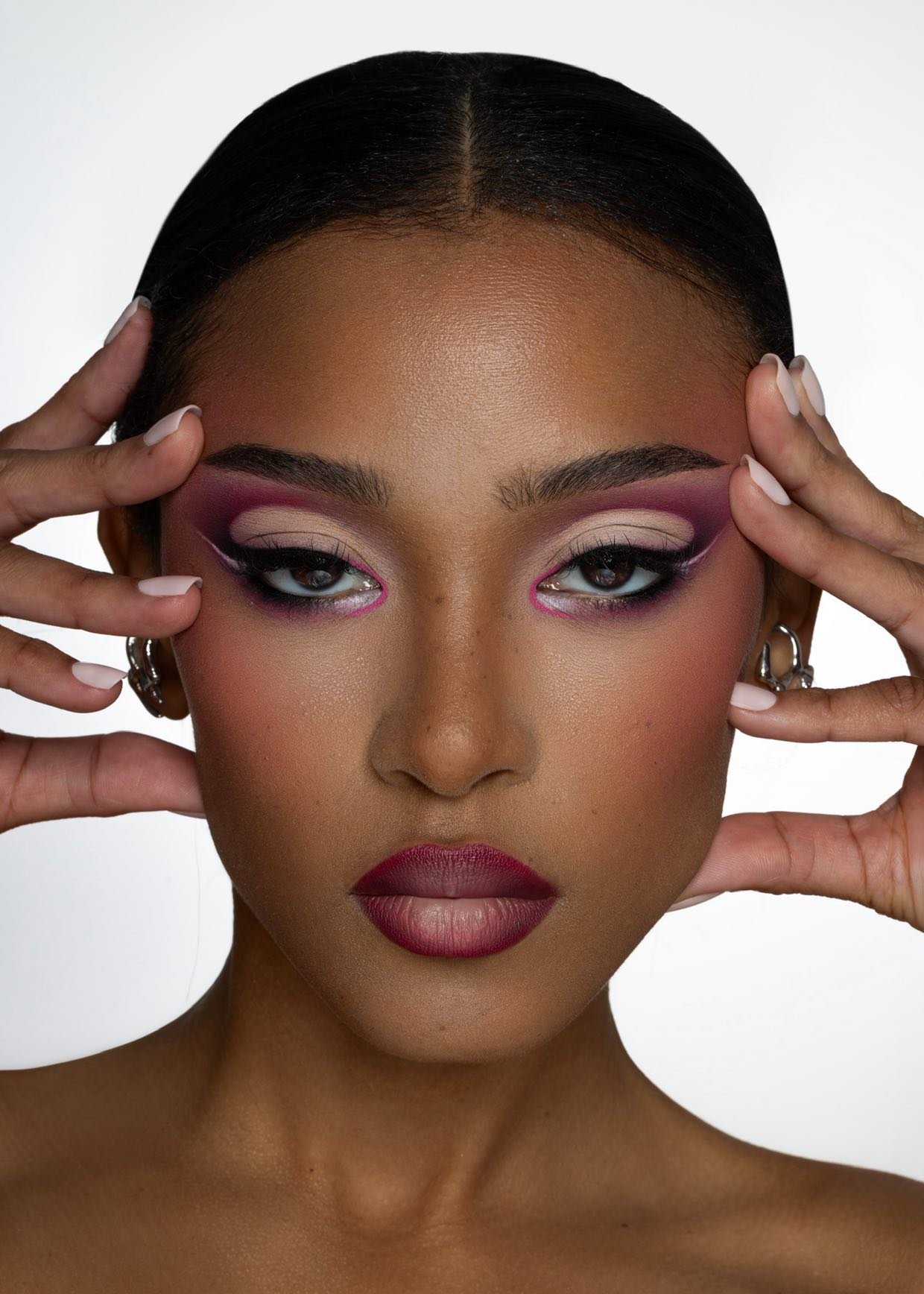 Close-up portrait of a woman with dark hair, bold makeup including pink, purple, and white eyeshadow, and dark lipstick. She is holding her face with both hands, showing her makeup and jewelry.