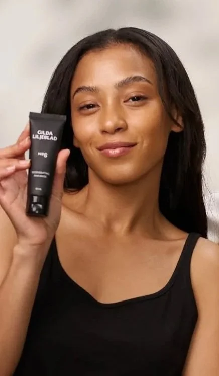 Young woman with dark hair holding a black tube of  skincare product, smiling at the camera.