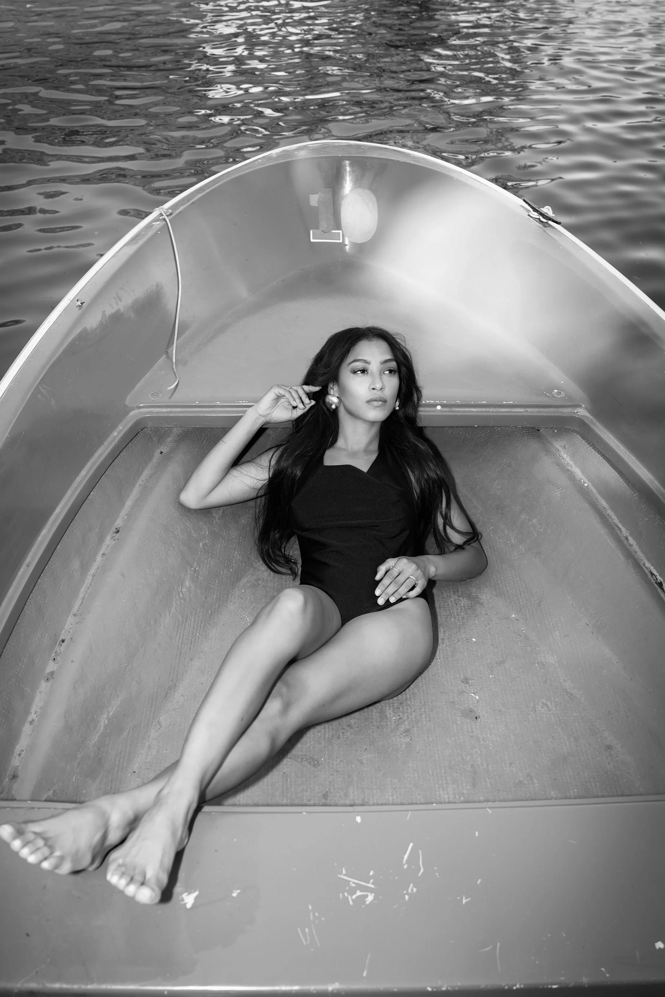 A woman lying down in a small boat on water, wearing a black dress, earrings, and with long hair, looking away from the camera.