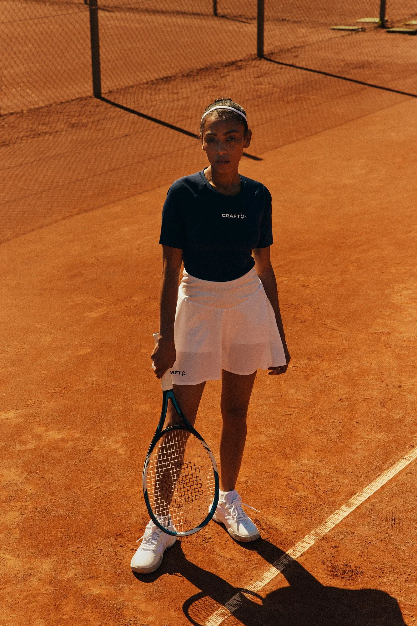 A woman standing on a clay tennis court holding a tennis racket, wearing a black top, white skirt, and white sneakers, with a tennis net in the background and sunlight casting shadows.