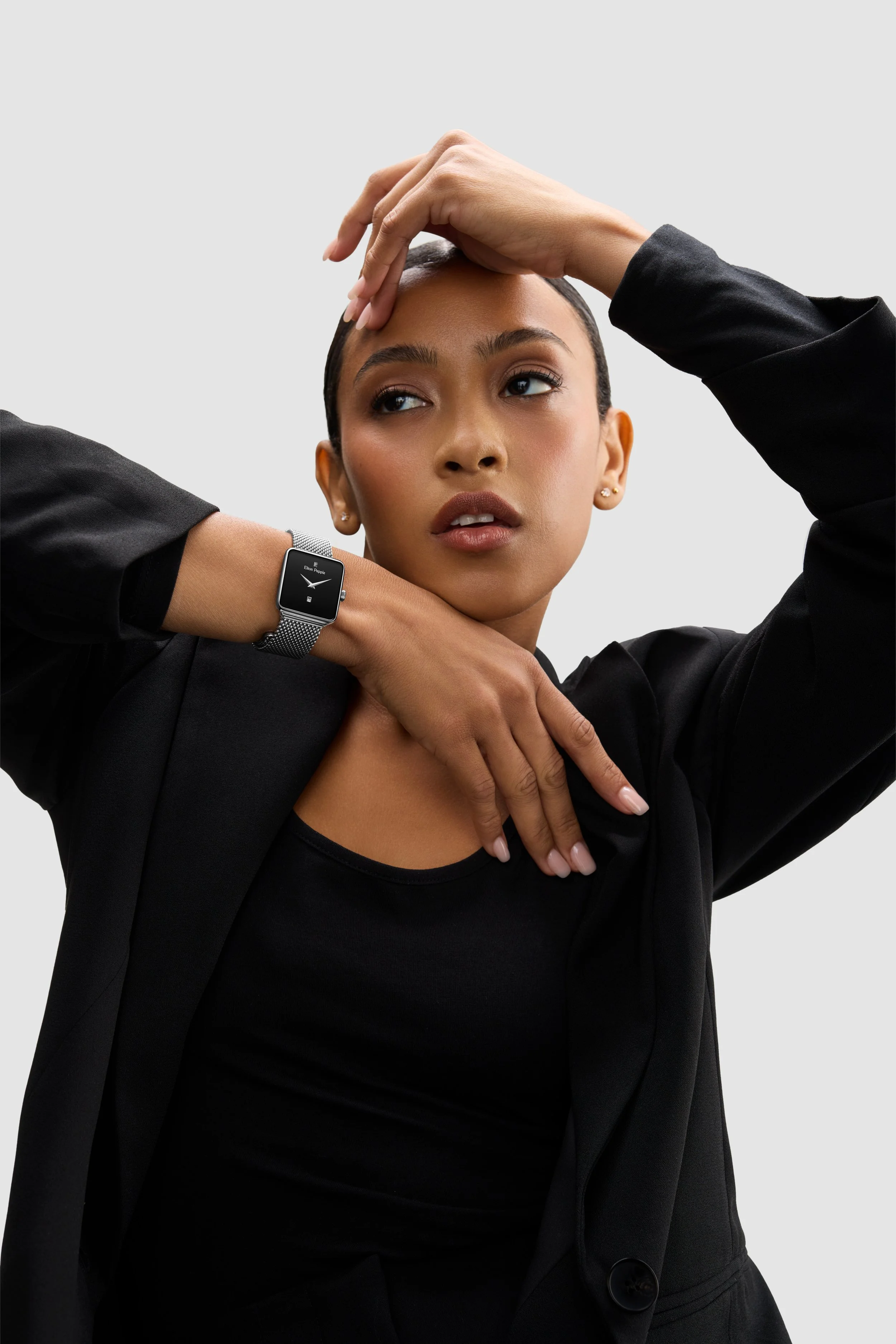 A woman with dark hair styled close to her scalp poses with one hand on her forehead and the other across her chest, wearing a black blazer and a smartwatch.