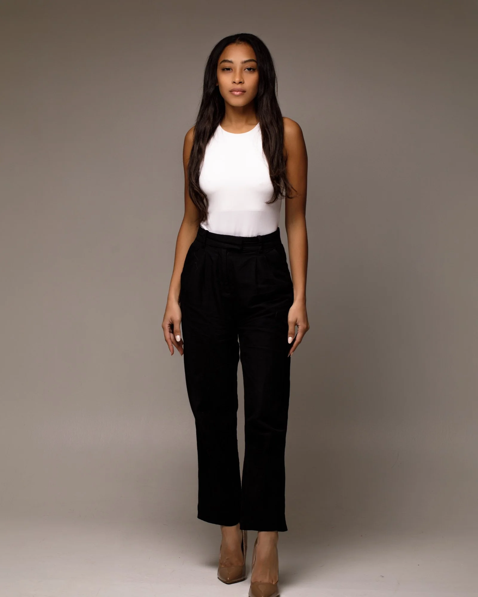 A woman standing against a plain gray background, wearing a white sleeveless top, black pants, and nude high heels.