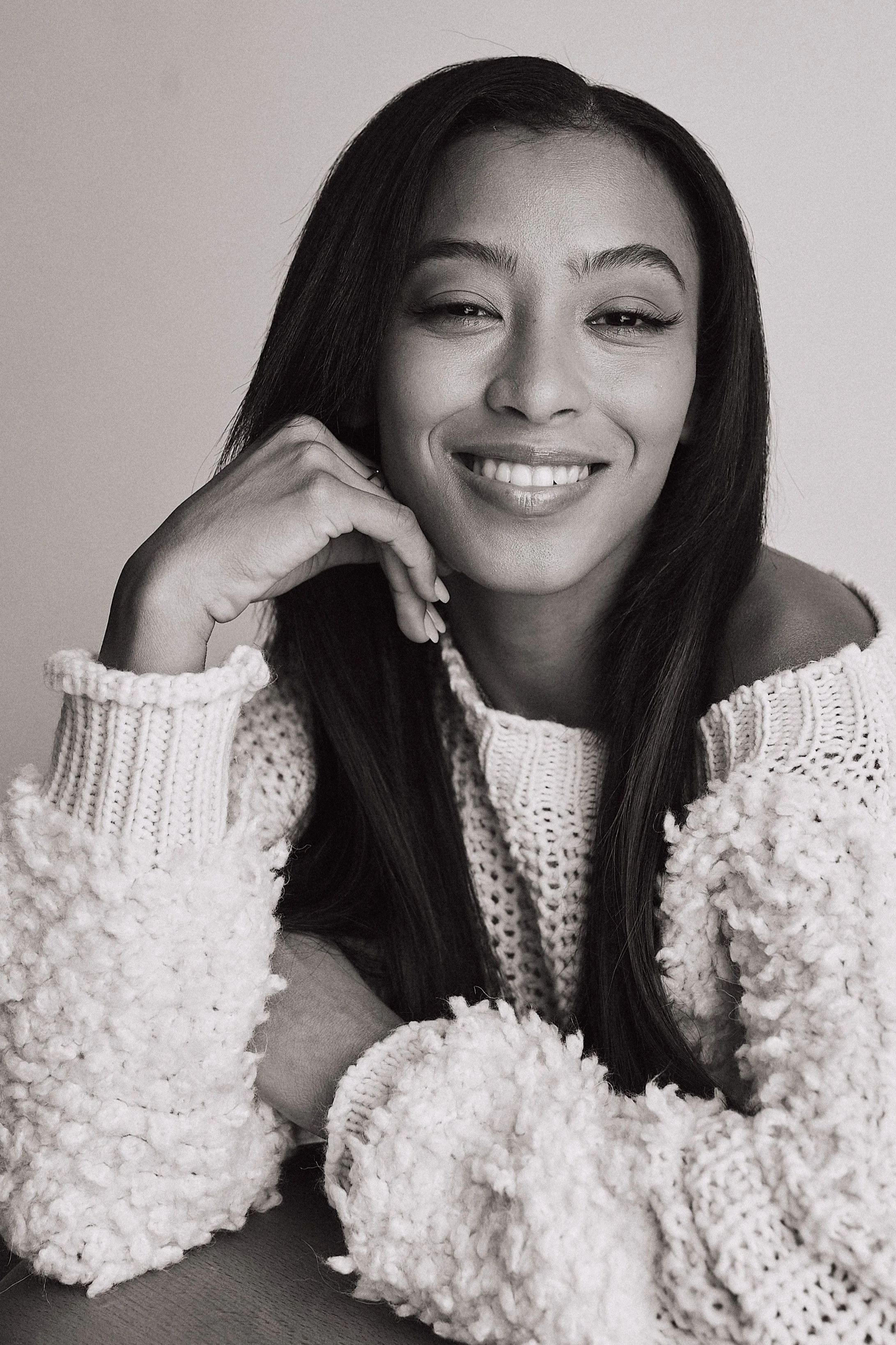 A woman with long dark hair smiling, resting her chin on her hand, wearing a cozy, textured sweater with large fluffy cuffs.