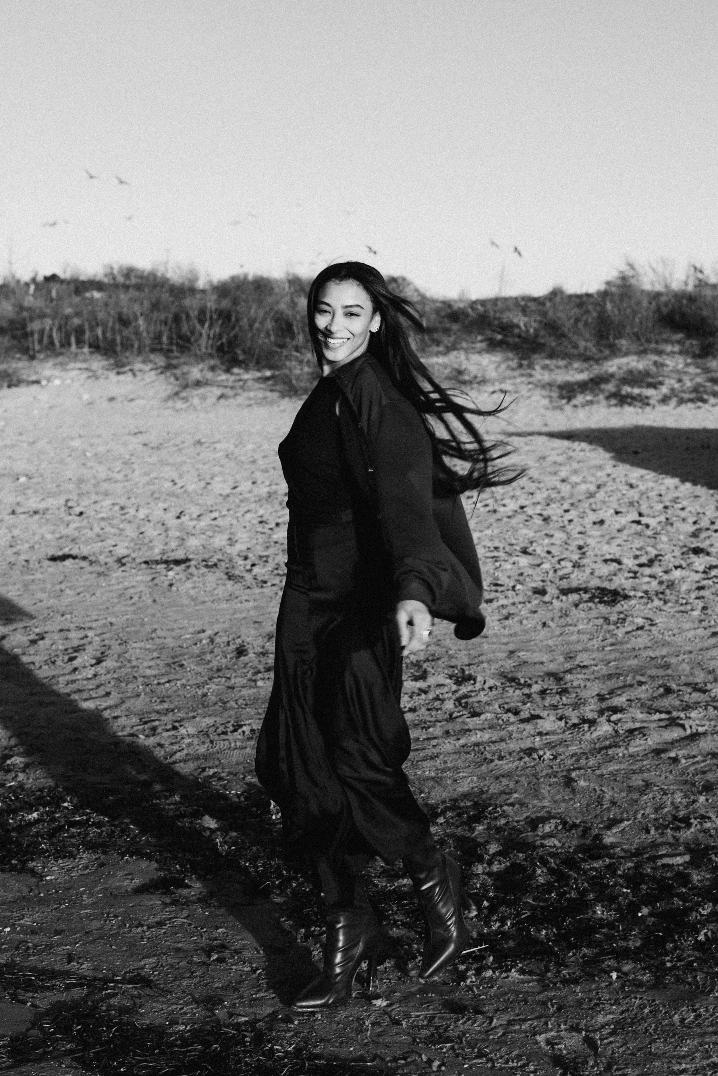 A woman smiling and winking, wearing a black outfit and high-heeled boots, standing on a beach with sand and sparse vegetation, with birds flying in the background.