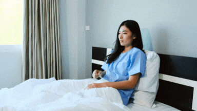A woman sitting in bed wearing a hospital gown, holding a cup, in a bright room with a window and curtains.