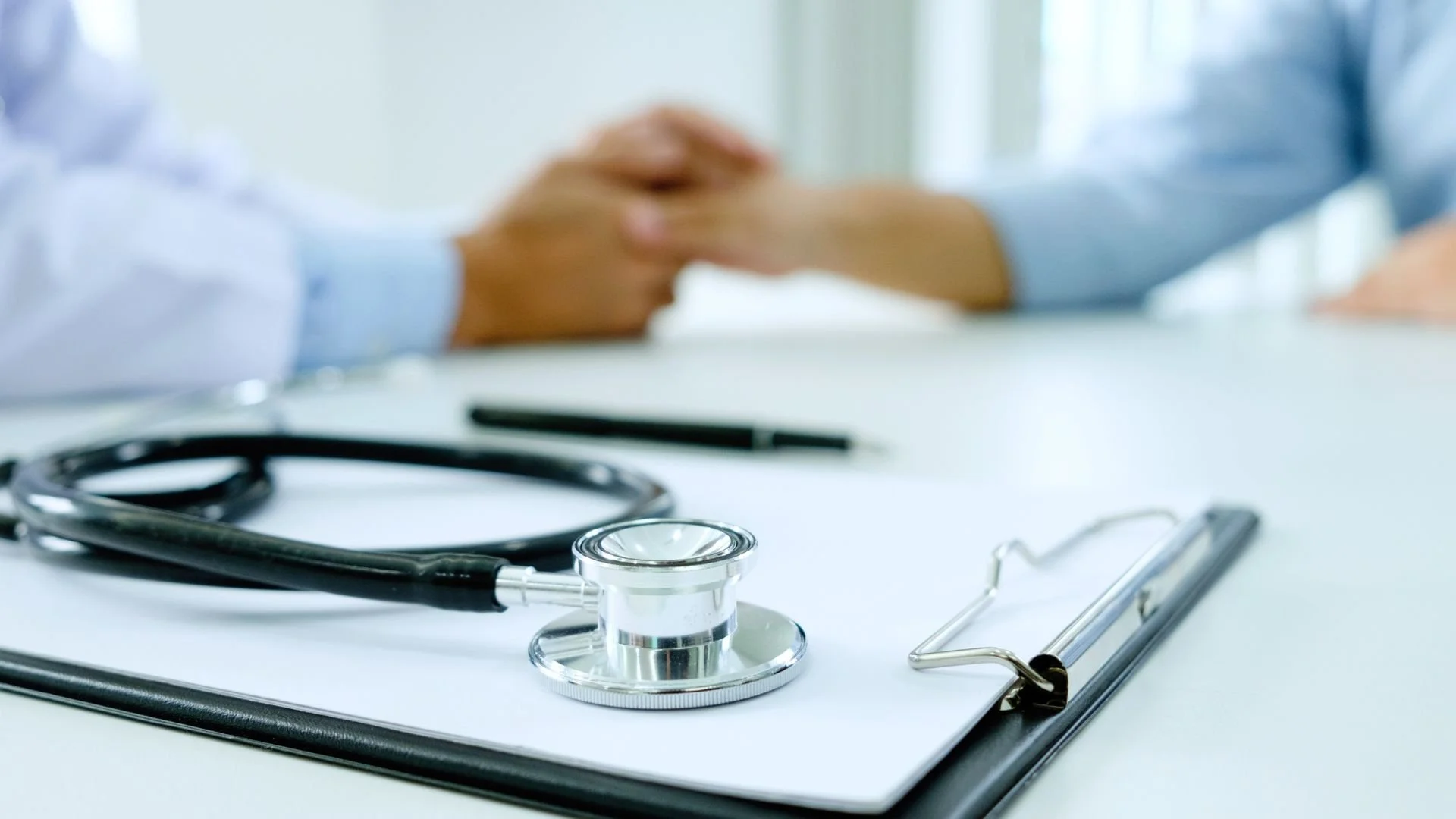 Close-up of a stethoscope on a clipboard with two people shaking hands in the background.