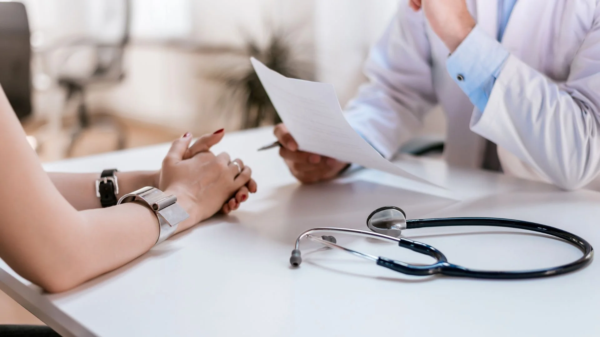 Doctor in white coat holding a paper while sitting at a desk with a patient, a stethoscope is placed on the desk.