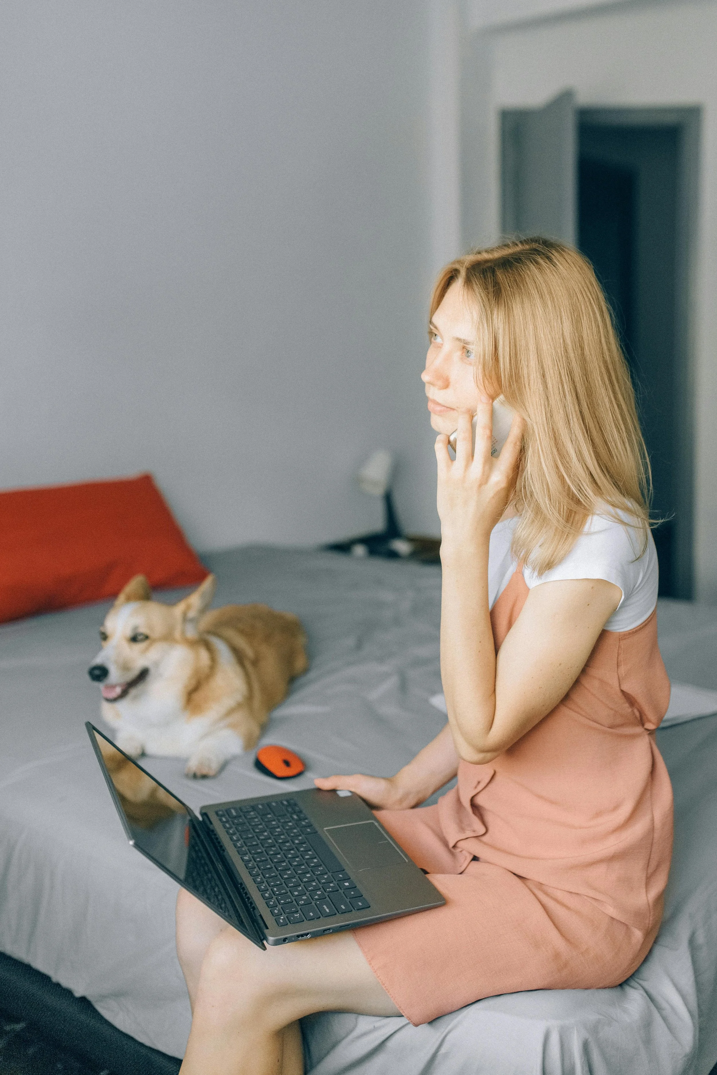 A woman sitting on a bed with a laptop on her lap, talking on her phone, with a Corgi dog lying on the bed nearby.