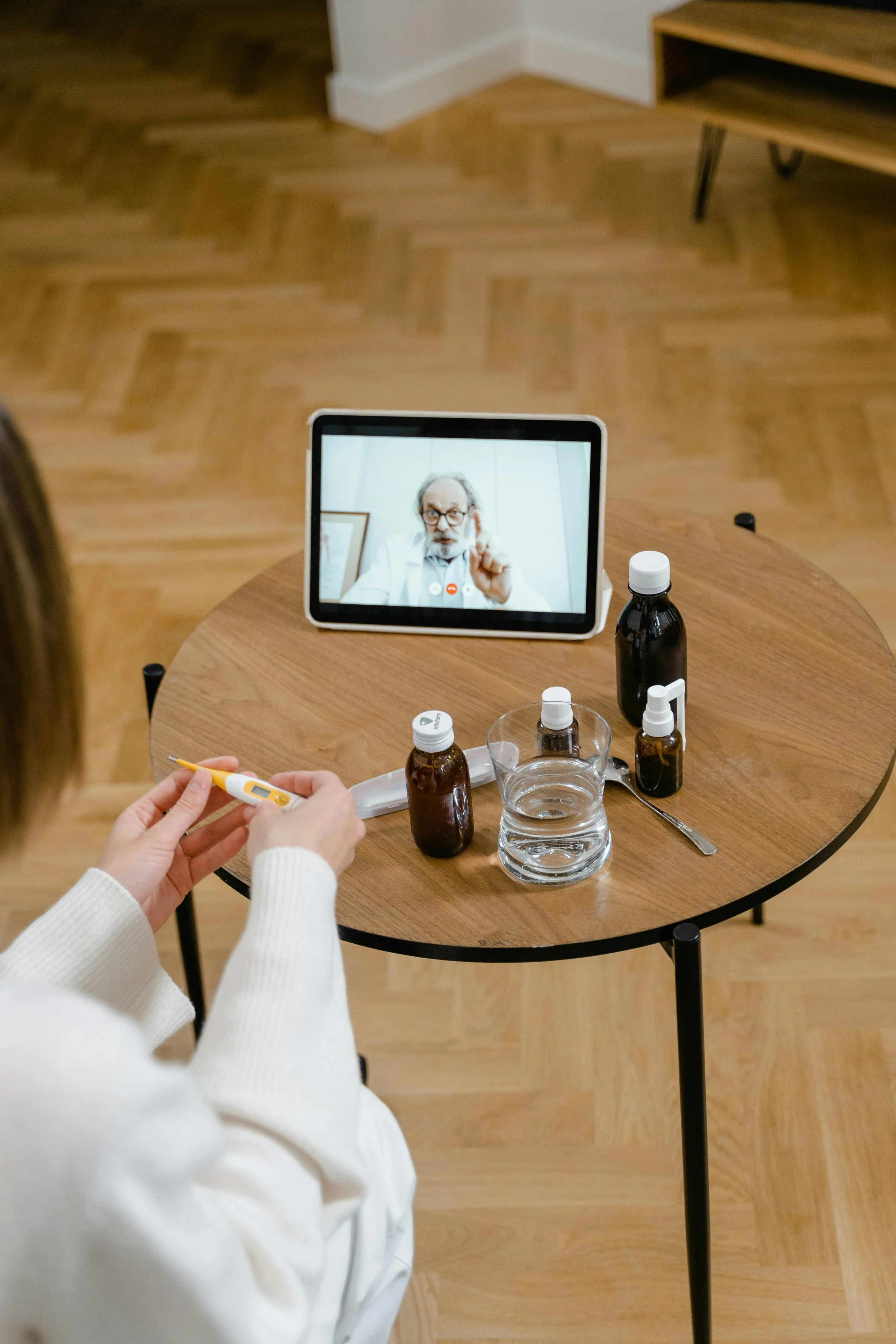 A woman holding a digital thermometer sitting at a wooden table with medicine bottles, a glass of water, and a spoon, during a video call with a doctor on a tablet. The doctor appears to be giving medical advice.
