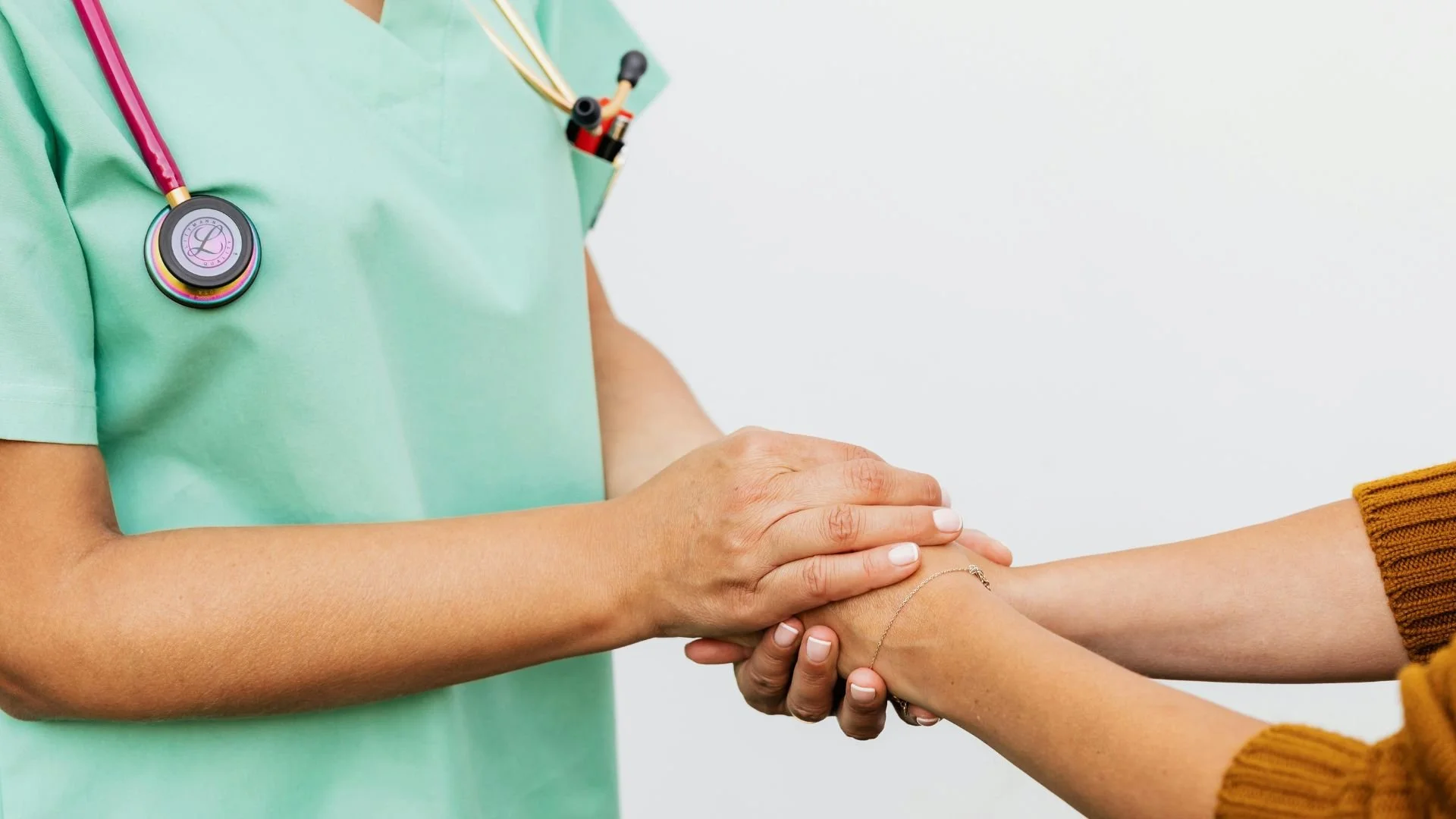 A healthcare professional, wearing a light green medical coat with a stethoscope around her neck, is holding hands with a patient. The patient is wearing a mustard yellow sweater. The background is plain white.