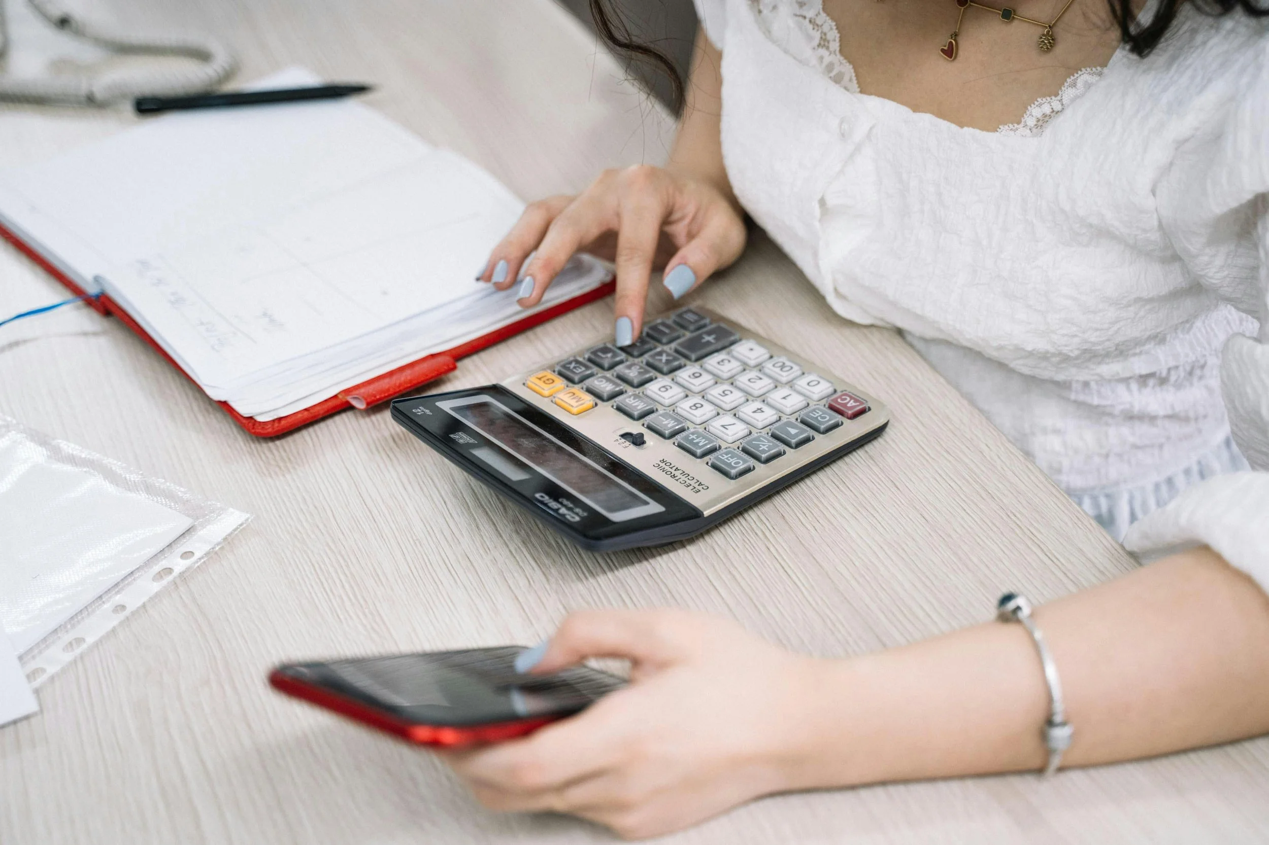 A woman sitting at a desk with a calculator, a red notebook, a pen, a smartphone, and some papers, appearing to work on finances or budgeting.