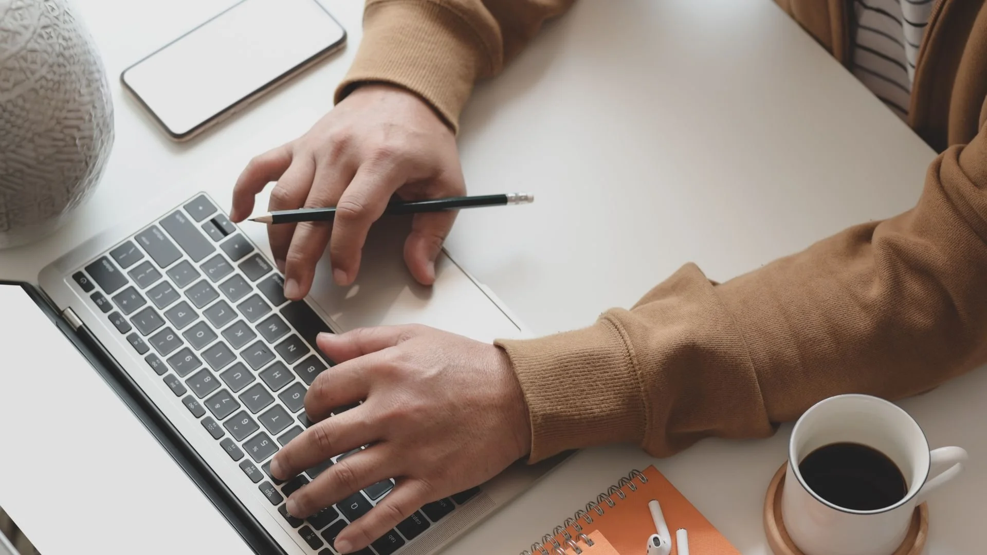 Person typing on a laptop keyboard with a pencil in hand, a coffee mug, a smartphone, and a notebook on a white desk.