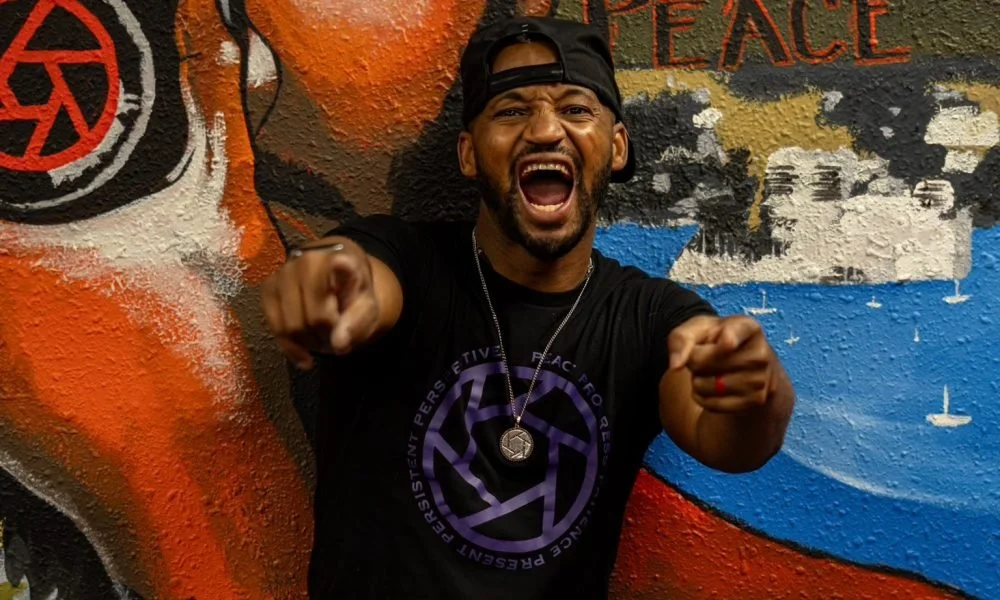 A man with a black baseball cap backwards, a black T-shirt with a purple logo, and a silver necklace, shouting and pointing towards the camera against a colorful graffiti wall.