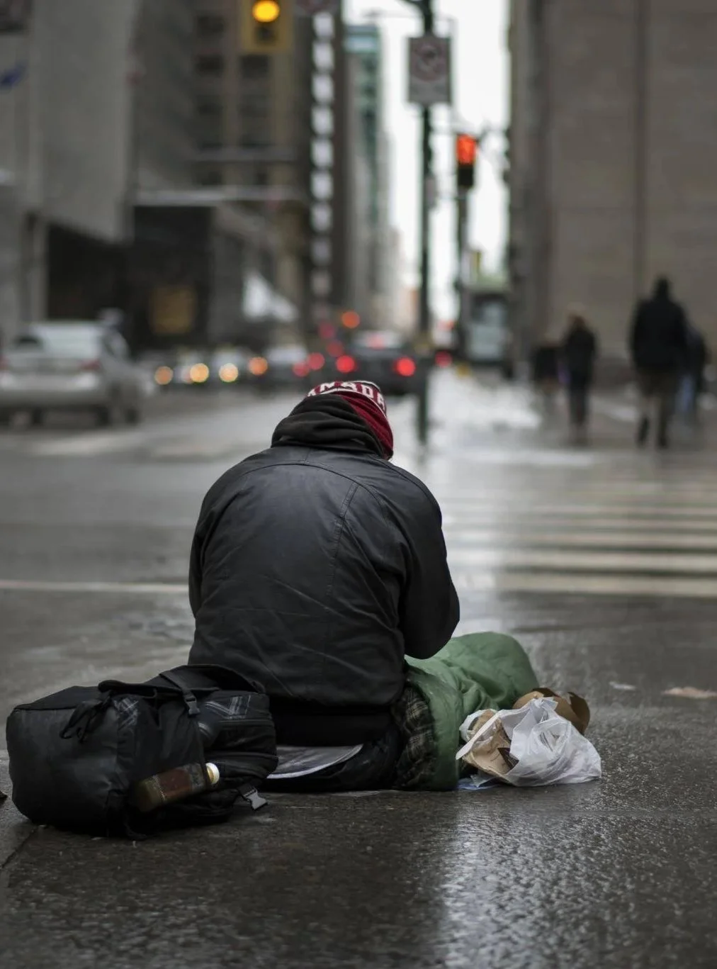 A homeless person sitting on the wet city street corner, wearing a black jacket and red beanie, surrounded by a backpack and plastic bags.