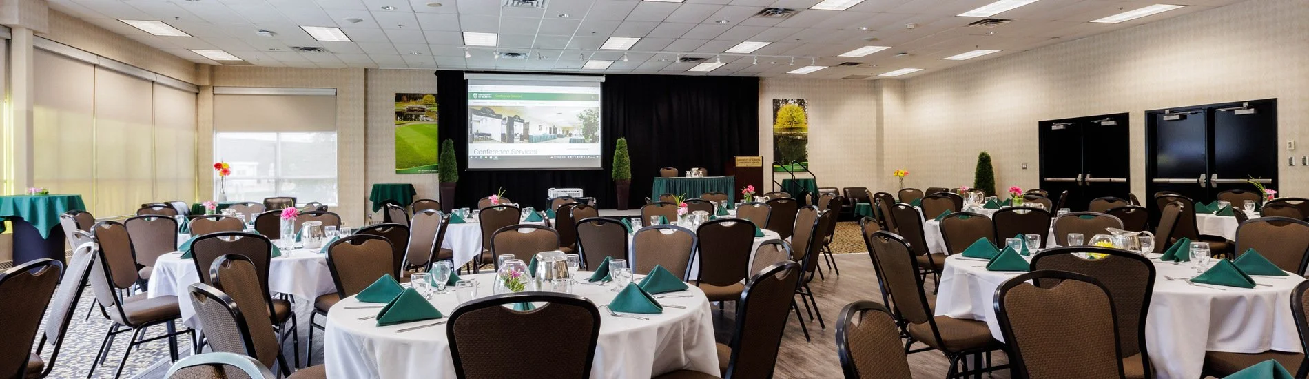Conference room set up for an event with round tables covered in white tablecloths, each with teal napkins, glasses, and small centerpieces with pink flowers. There is a large projection screen at the front displaying a presentation, with black curtains behind it. The room has beige walls, large windows with blinds, and a carpeted floor.