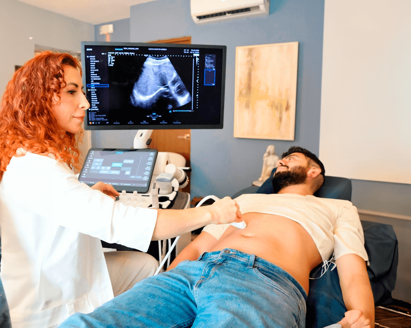 A woman is performing an ultrasound scan on a man lying on a medical examination table. The woman is holding a transducer against the man's abdomen, and an ultrasound monitor displays a black-and-white image of internal organs. The room has blue walls, a framed art piece, and an air conditioning unit.