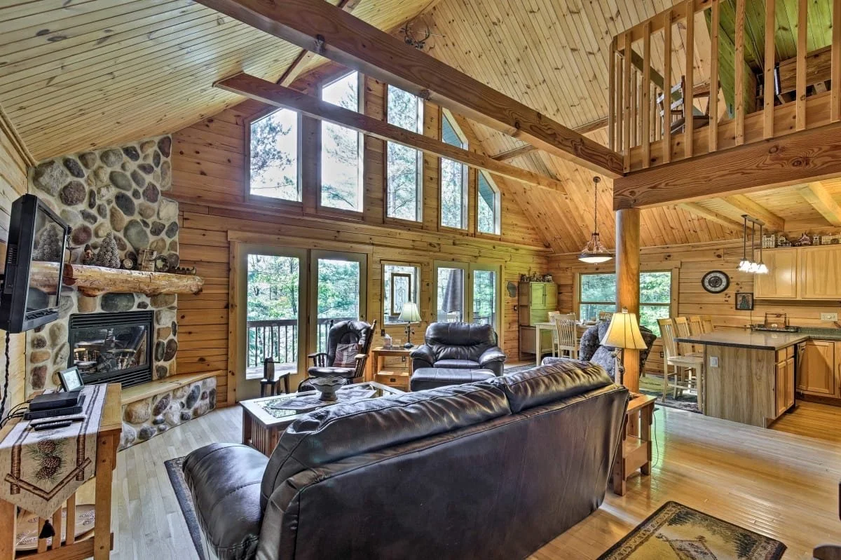 Interior view of a cozy wood-paneled living room with high vaulted ceilings, large windows, and a stone fireplace. The room features leather furniture, a wooden coffee table, and a kitchen area with a breakfast bar and dining table.