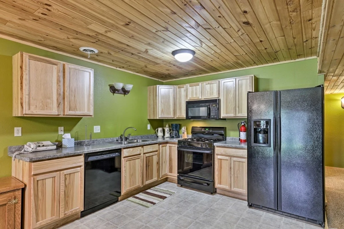 Kitchen with wooden cabinets, black refrigerator, microwave, stove, sink, green walls, wooden ceiling, and various small appliances.