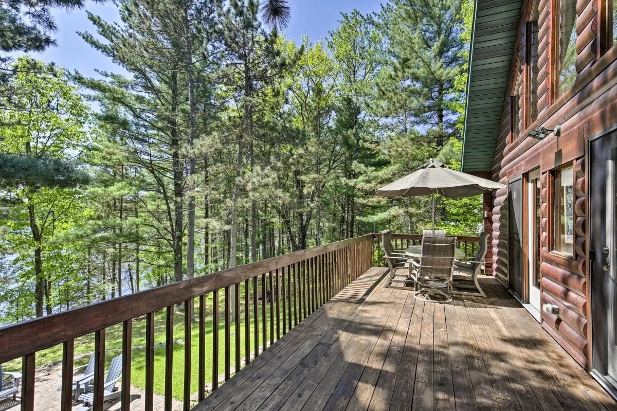 Wooden deck with outdoor dining table and chairs, umbrella, and view of a lush green forest.