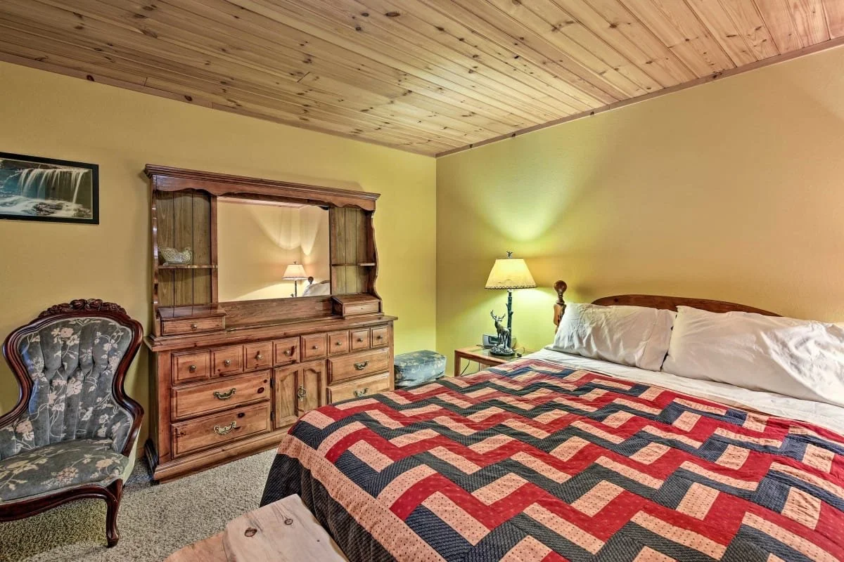 A cozy bedroom with a king-sized bed covered with a red, black, and beige quilt, a wooden headboard, two white pillows, a nightstand with a lamp and decorative figurine, an antique dresser with a mirror, a floral armchair, and a painted landscape pic