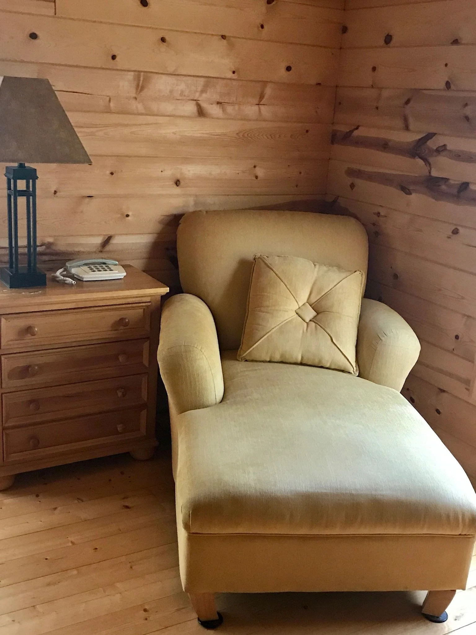 A cozy corner with a yellow armchair and a matching pillow, placed next to a wooden nightstand with a lamp and phone, in a room with wood-paneled walls and floor.
